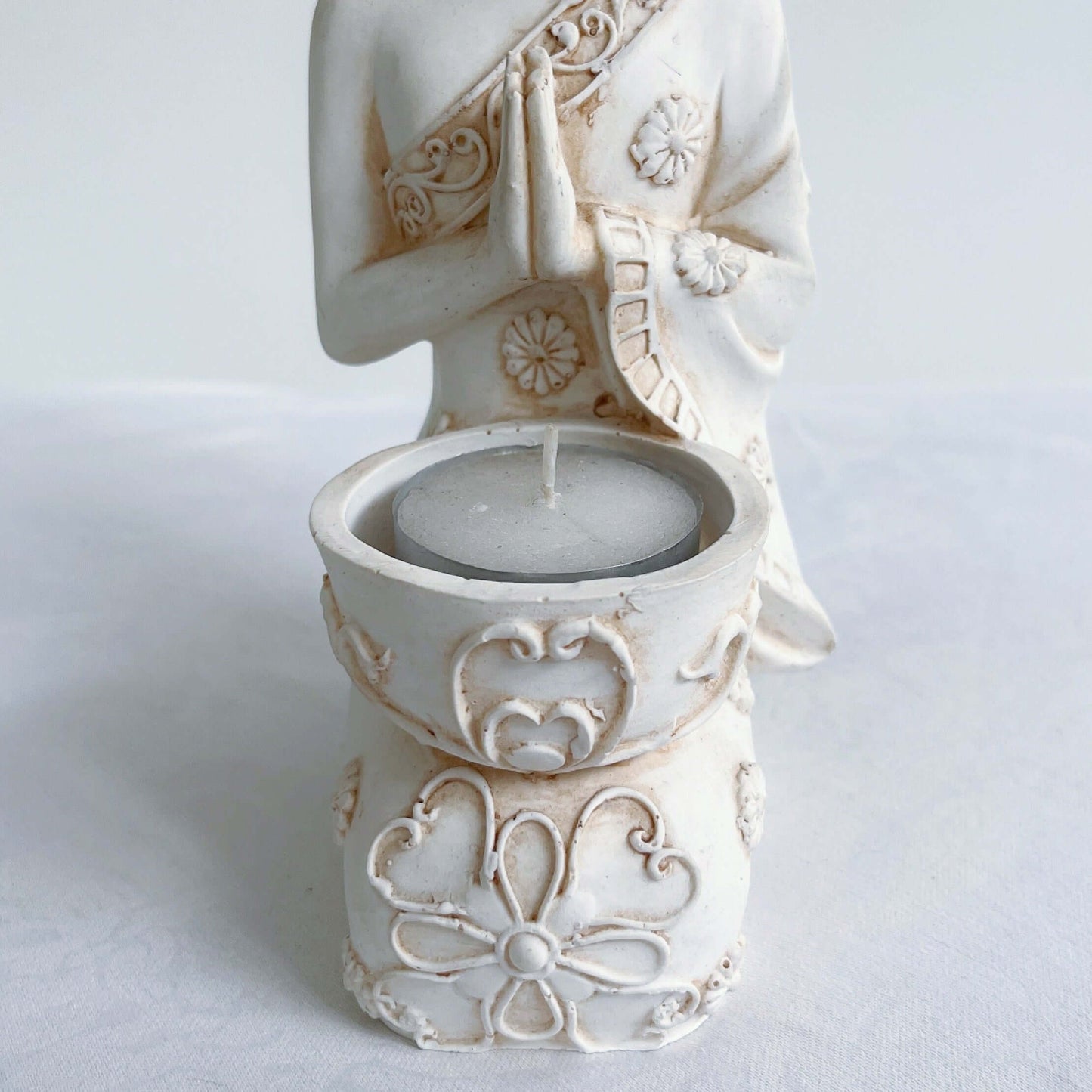 Detail view of buddha kneeling, candle holder on lap, with ornate robes and detailing, natural off white colors.