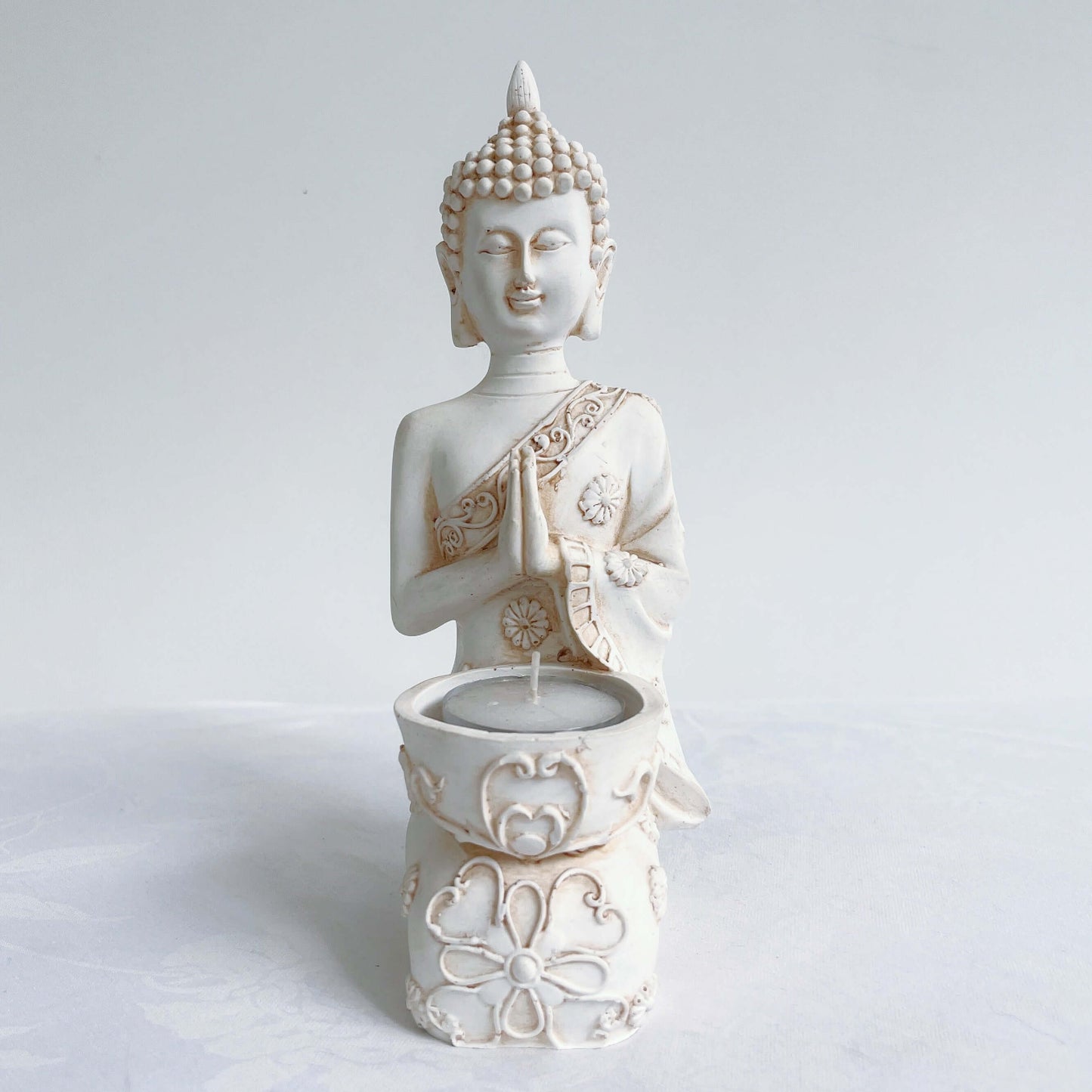 Front view of buddha kneeling, candle holder on lap, with ornate robes and detailing, natural off white colors.