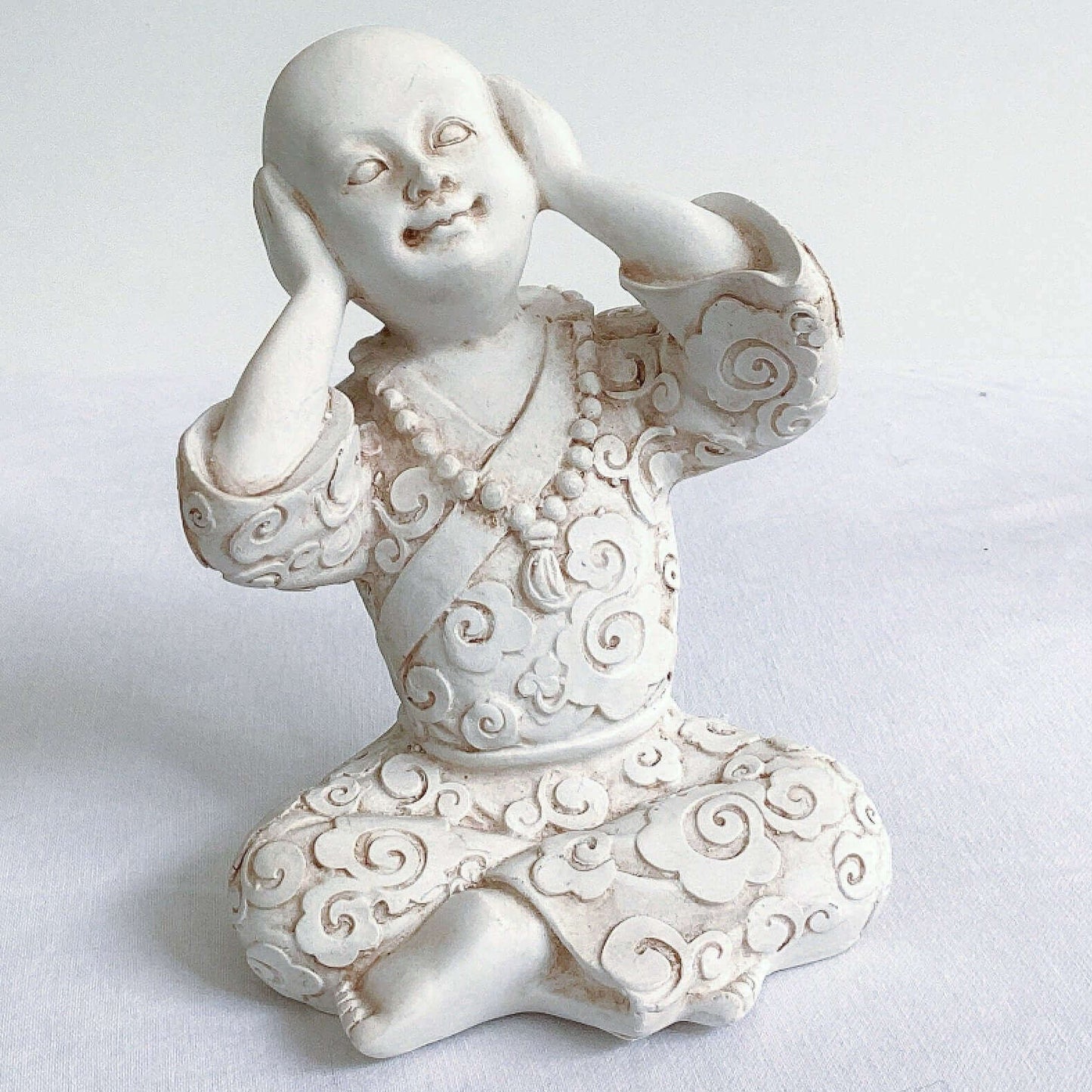 View of single monk, hear no evil, natural off white colors, with ornate robes, and hands covering the ears.