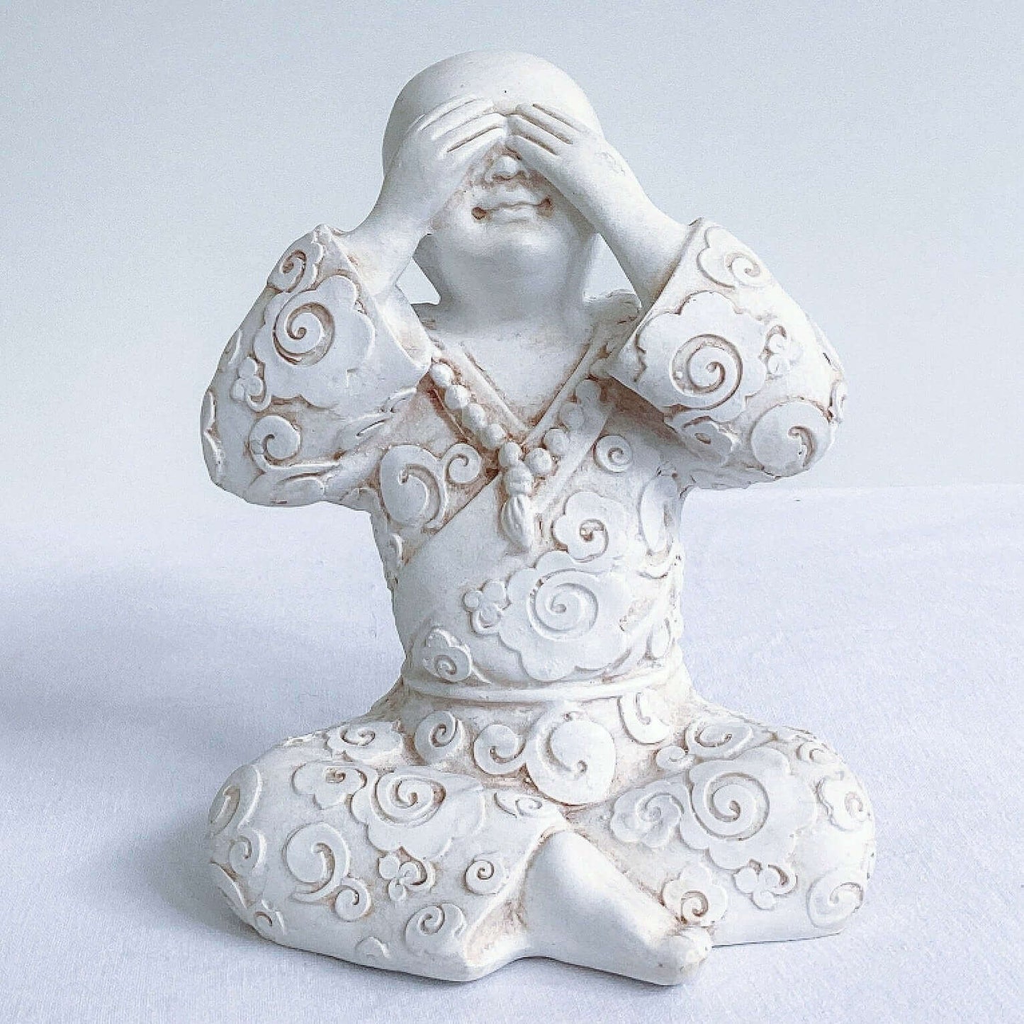 View of single monk, see no evil, natural off white colors, with ornate robes, and hands covering the eyes.