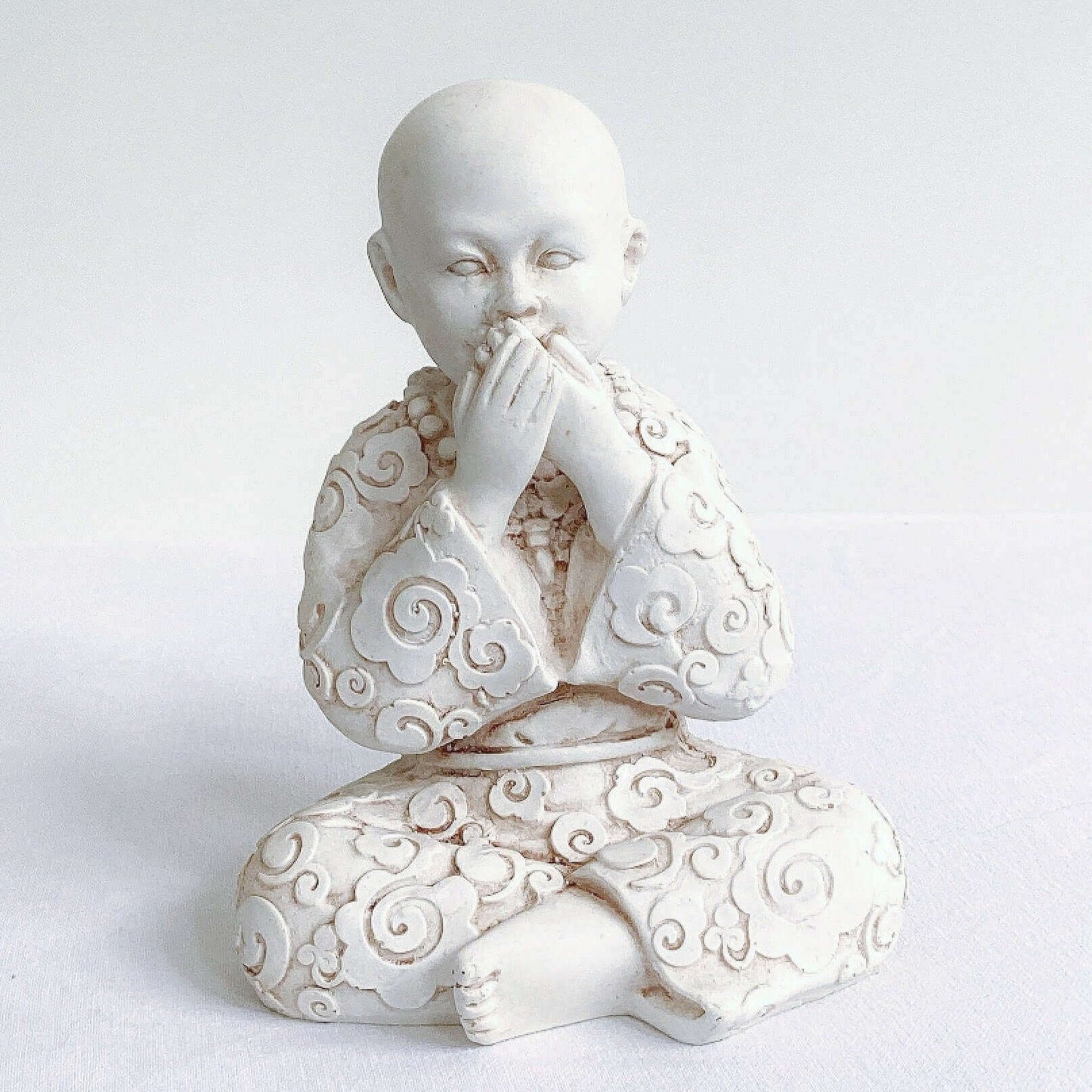 View of single monk, speak no evil, natural off white colors, with ornate robes, and hands covering the mouth.