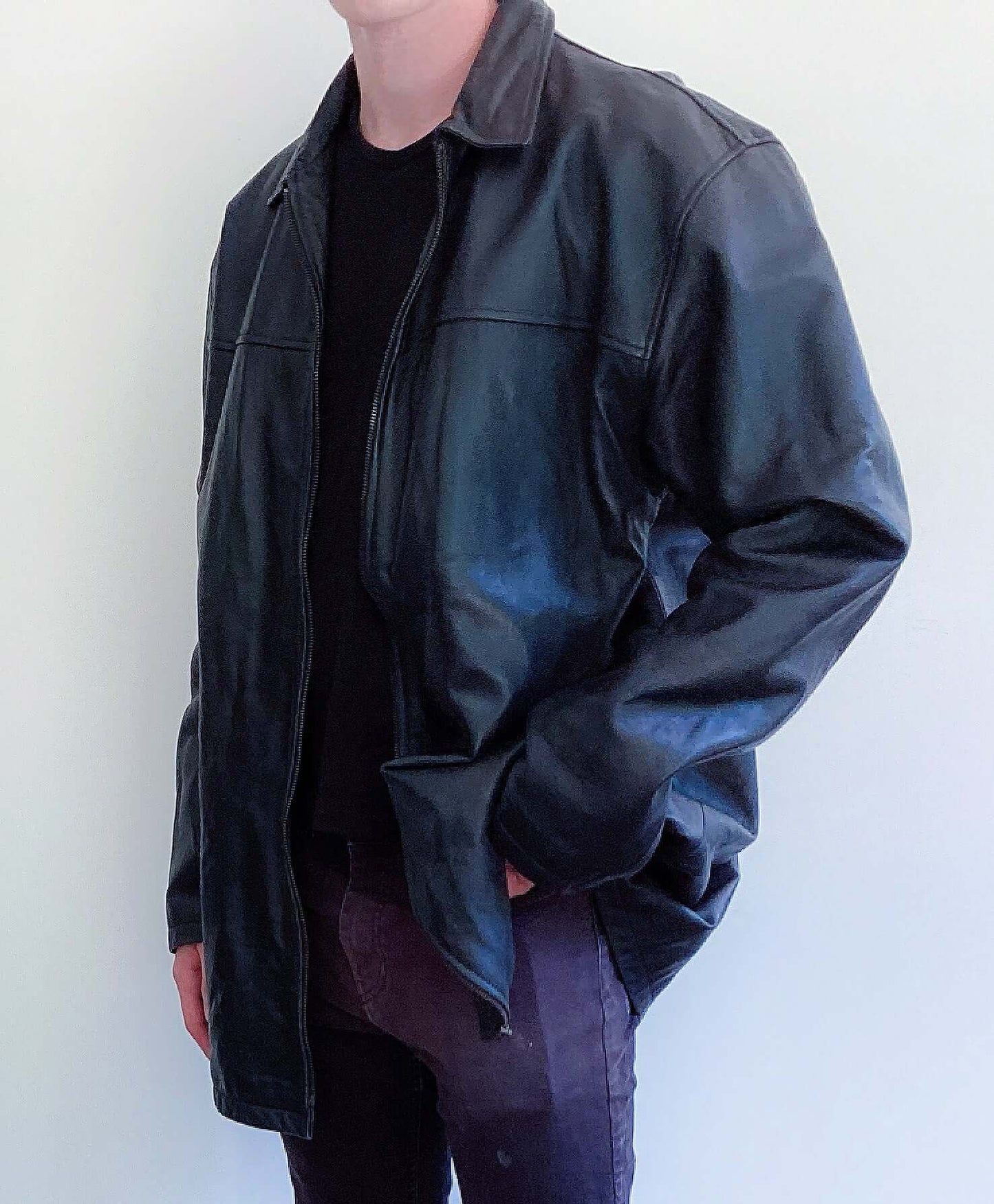 Side view of black leather jacket, mid thigh length, zip up, hands in pockets.