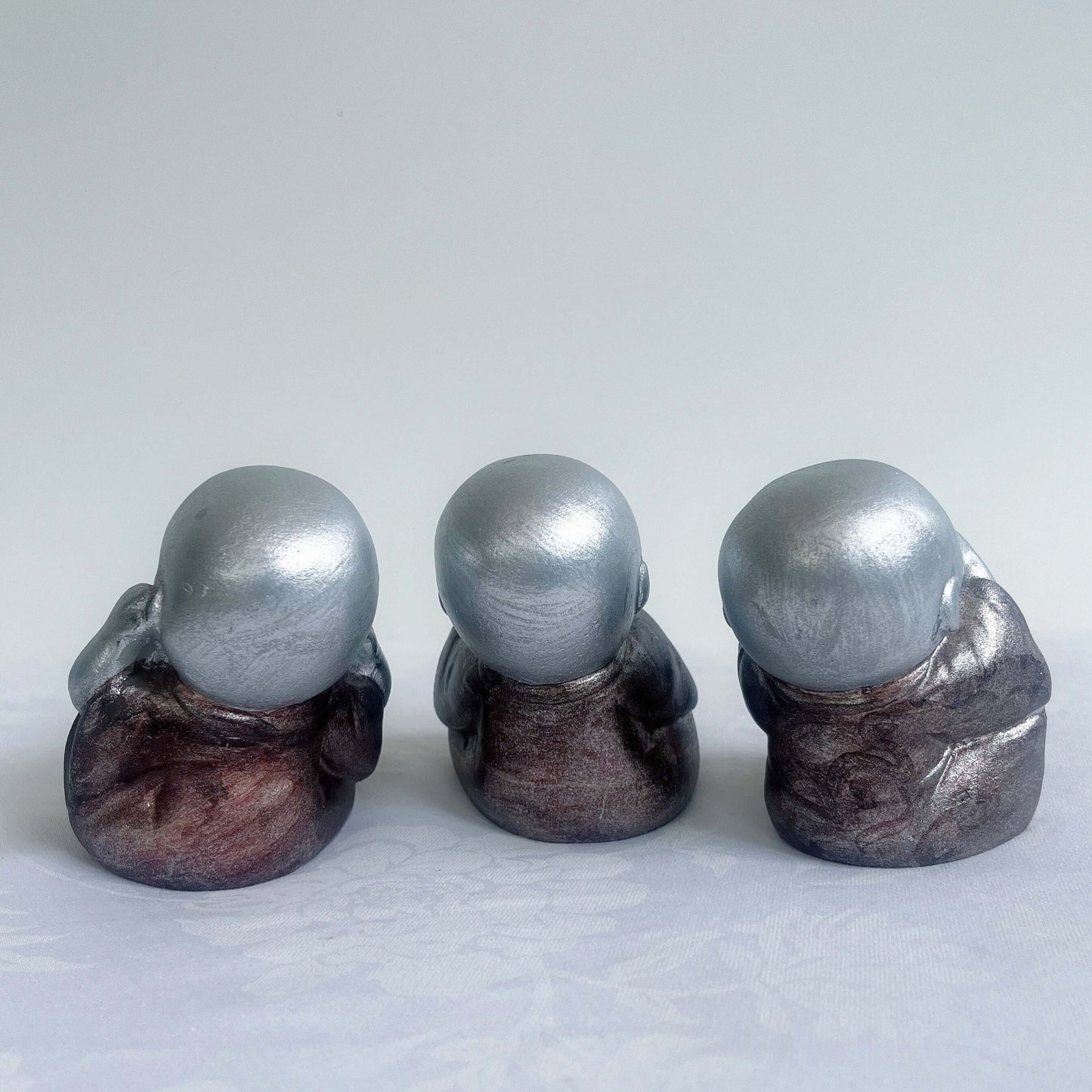 Back view of monk trio, painted silver figures with bronze robes, all sitting in a row.