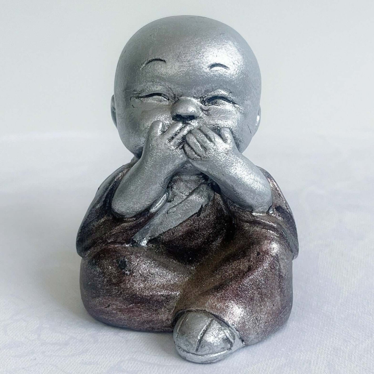 View of single monk, speak no evil, painted silver figure with bronze robes, and hands covering the mouth.
