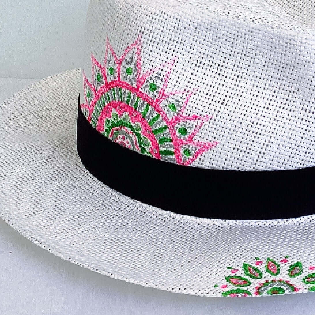 Detail view of panama hat, black band at base, with hand painted green and pink mandalas asymmetrically positioned.