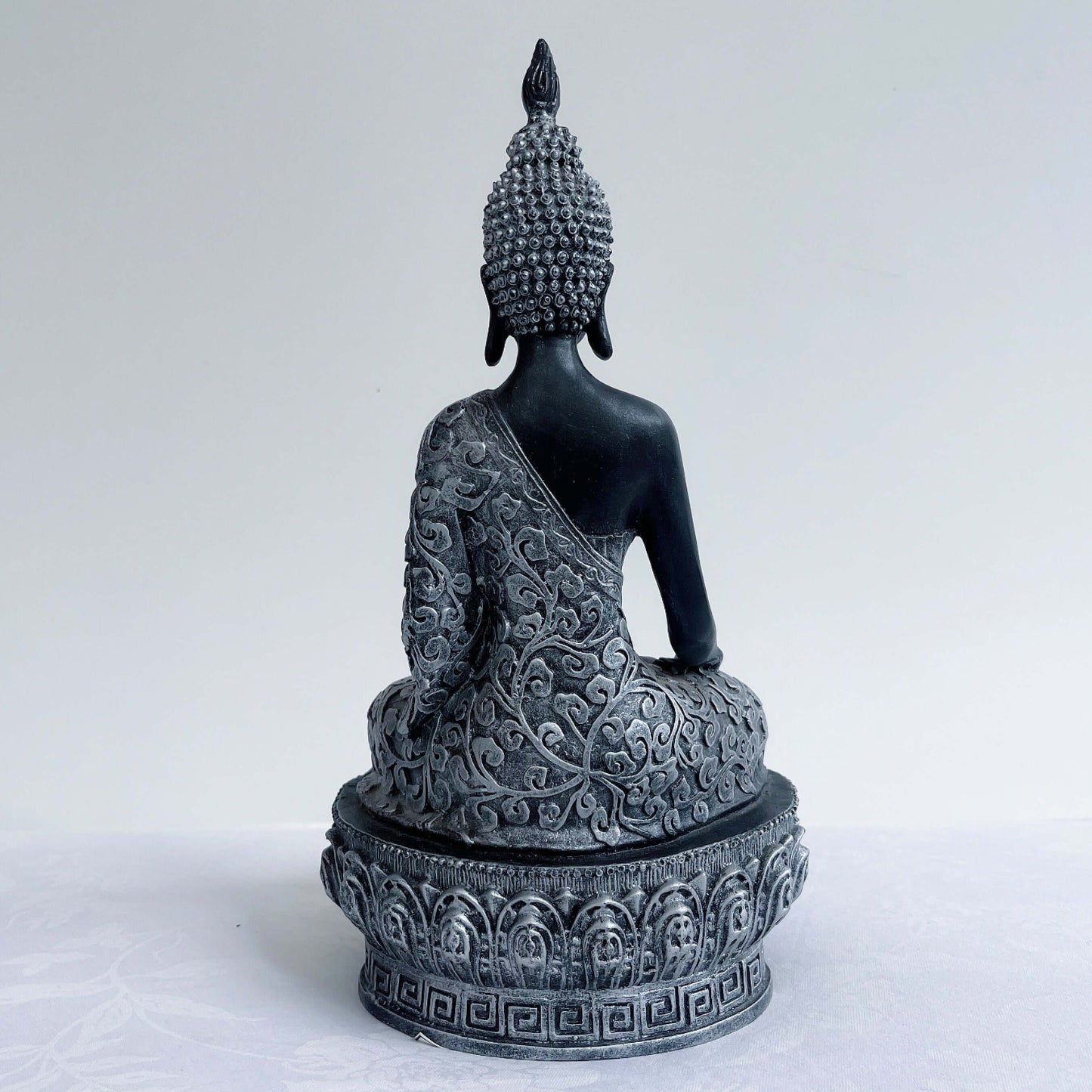 Back view of buddha in lotus position, black and silver paint wash, ornate robes and pedestal, ushnisha on head.