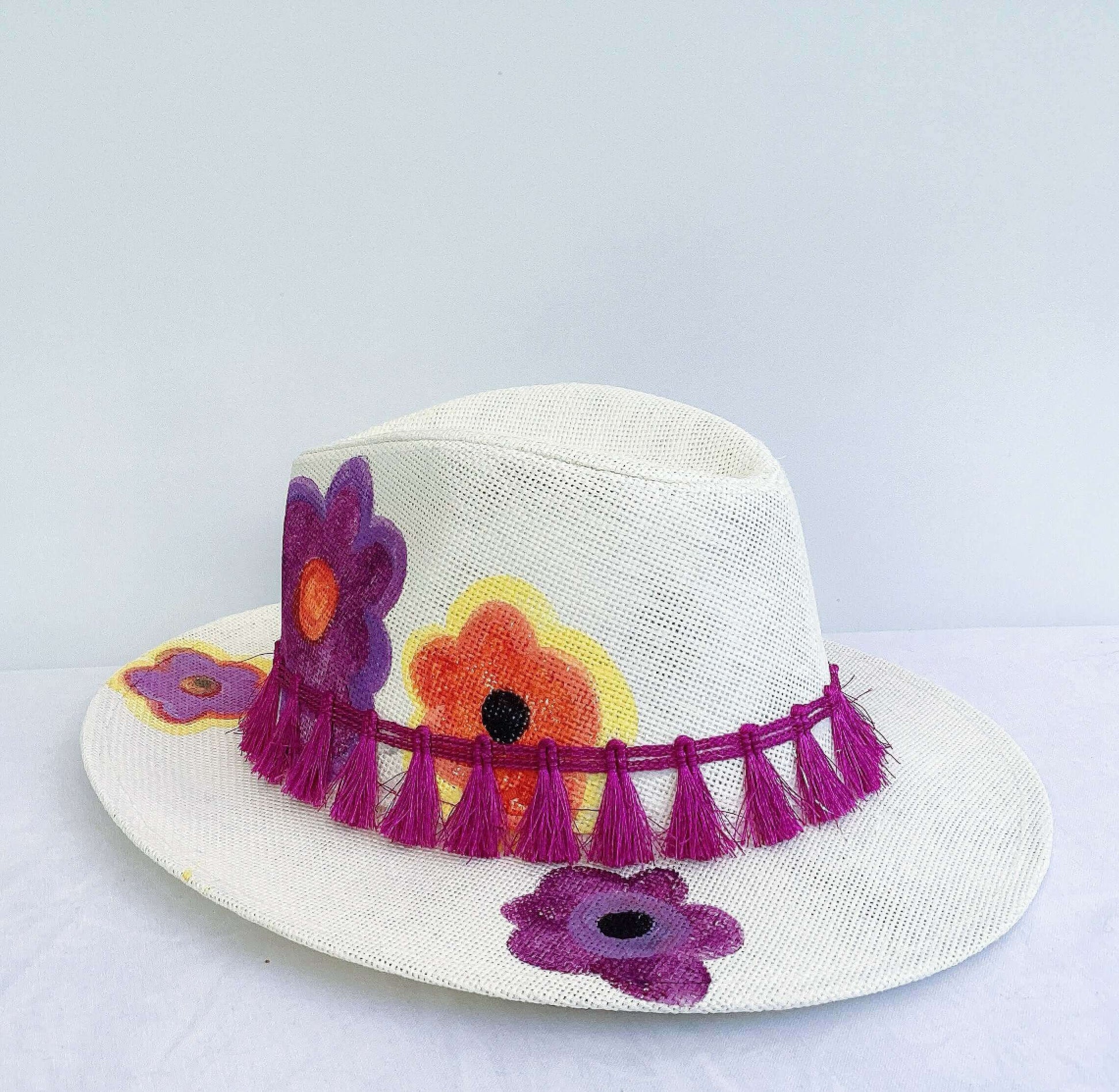 Side view of panama hat, black band and magenta fringe at base, painted purple and orange flowers asymmetrically spaced.