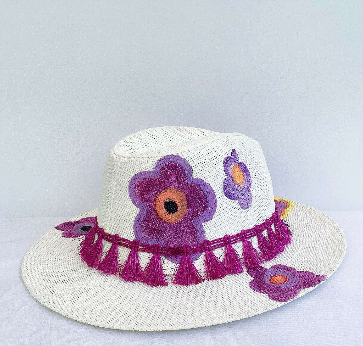 Detail view of panama hat, black band and magenta fringe at base, painted purple and orange flowers asymmetrically spaced.