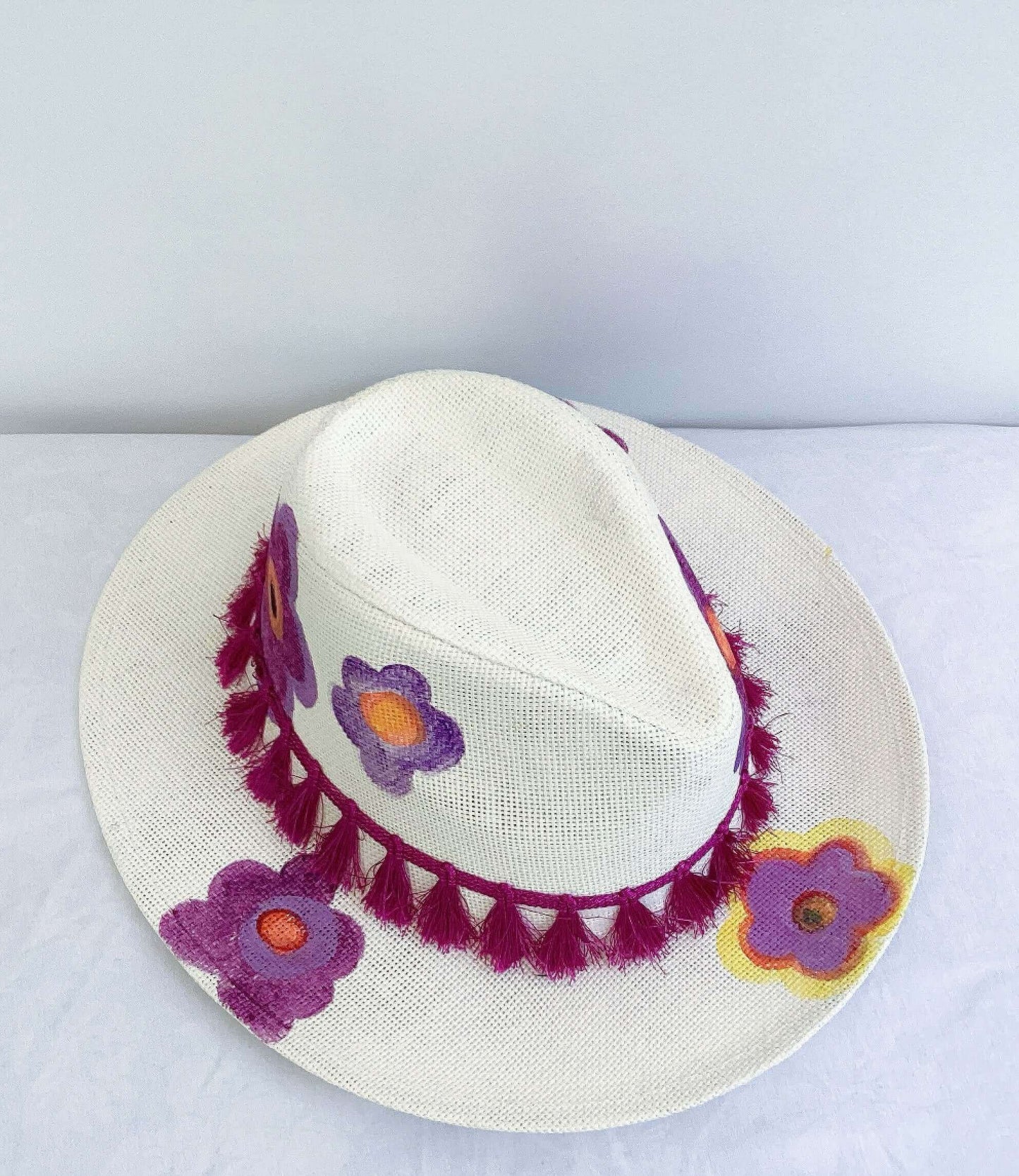 Front view of panama hat, black band and magenta fringe at base, painted purple and orange flowers asymmetrically spaced.