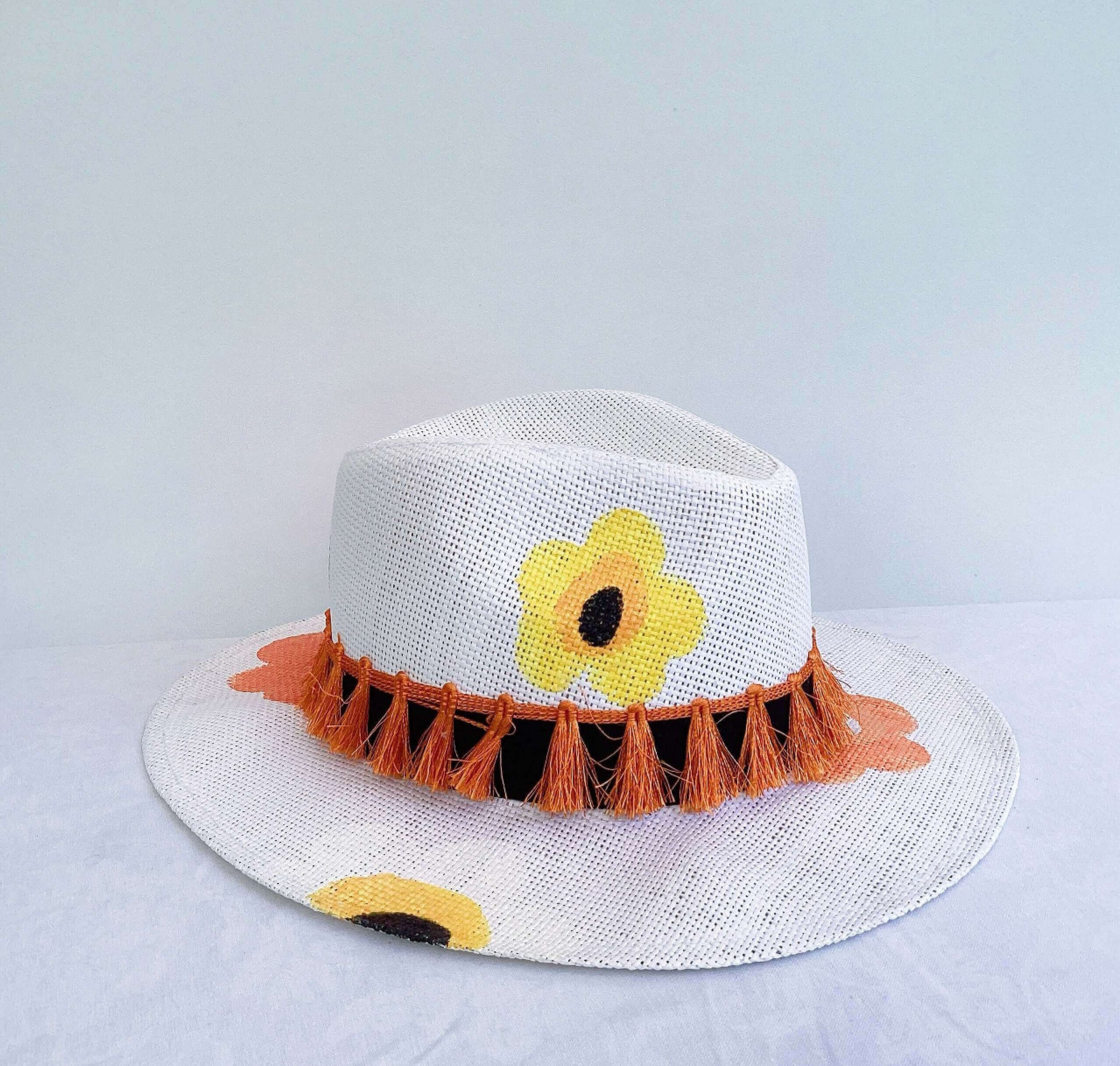 Detail view of panama hat, black band and orange fringe at base, painted yellow and orange flowers asymmetrically spaced.