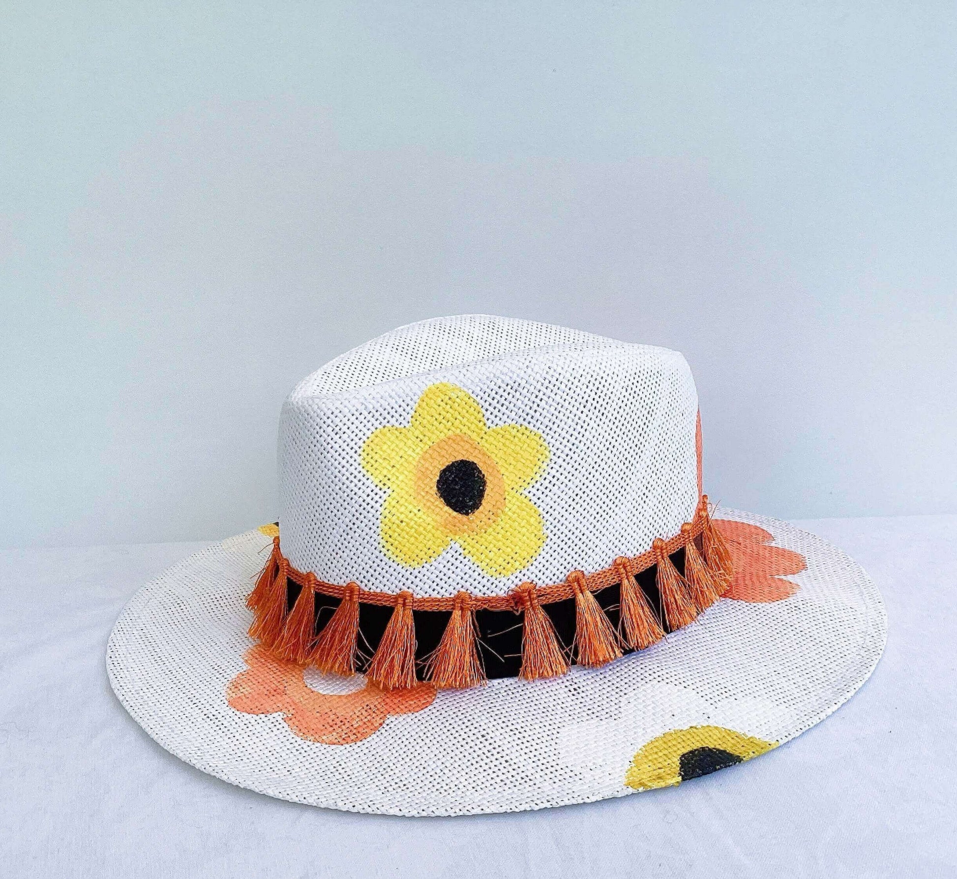 Front view of panama hat, black band and orange fringe at base, painted yellow and orange flowers asymmetrically spaced.