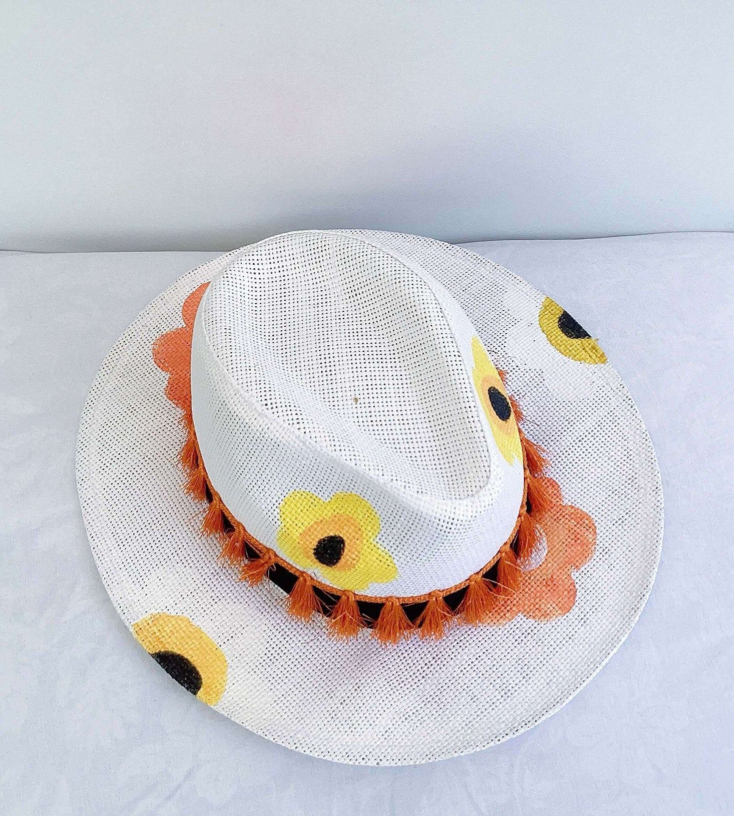 Side view of panama hat, black band and orange fringe at base, painted yellow and orange flowers asymmetrically spaced.