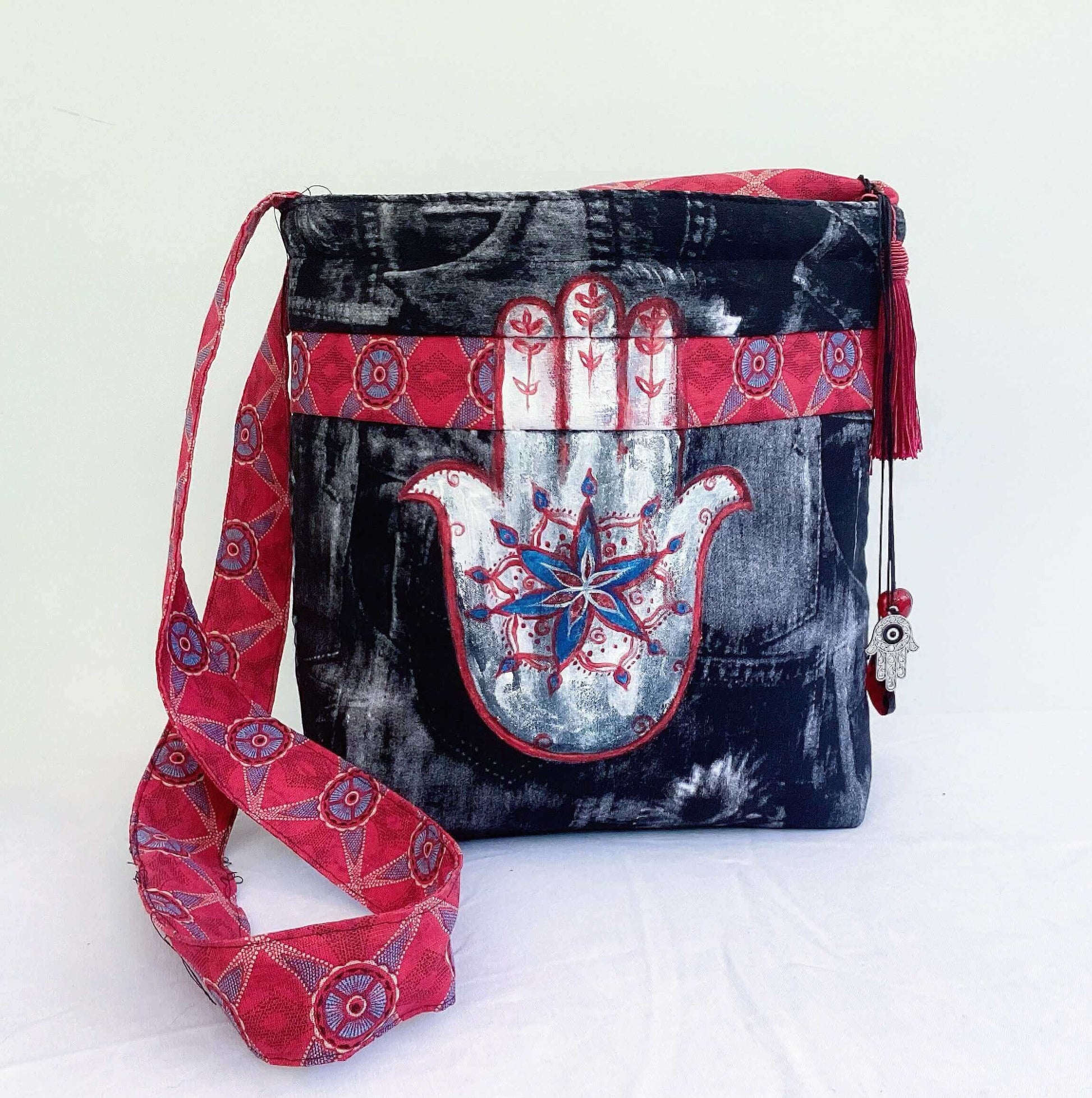 Front of bag, black and white fabric, white, red and blue painted hamsa, beads hanging off the red shwe shwe fabric strap.