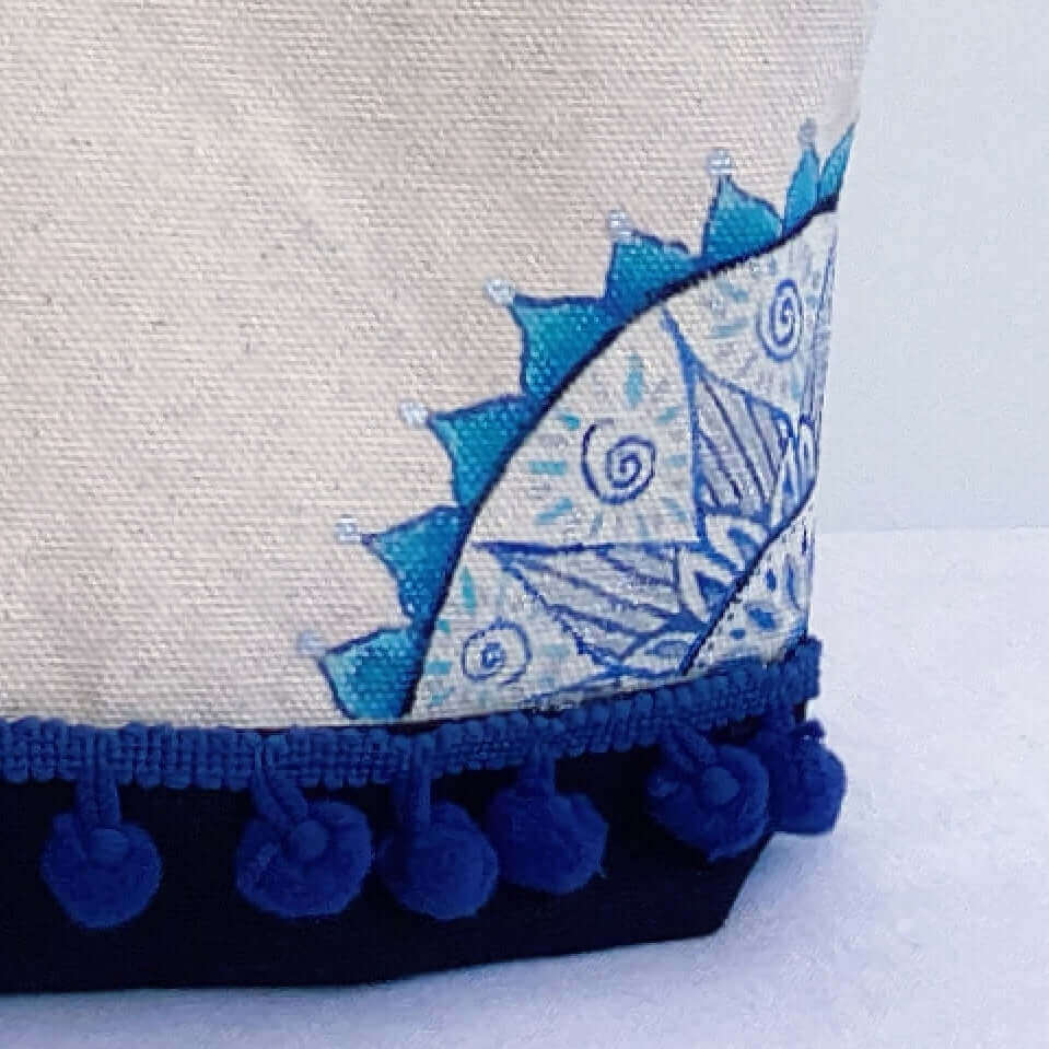 Detail view of canvas make-up pouch, black canvas base, with hand painted blue mandalas and zip, blue pom pom fringe.