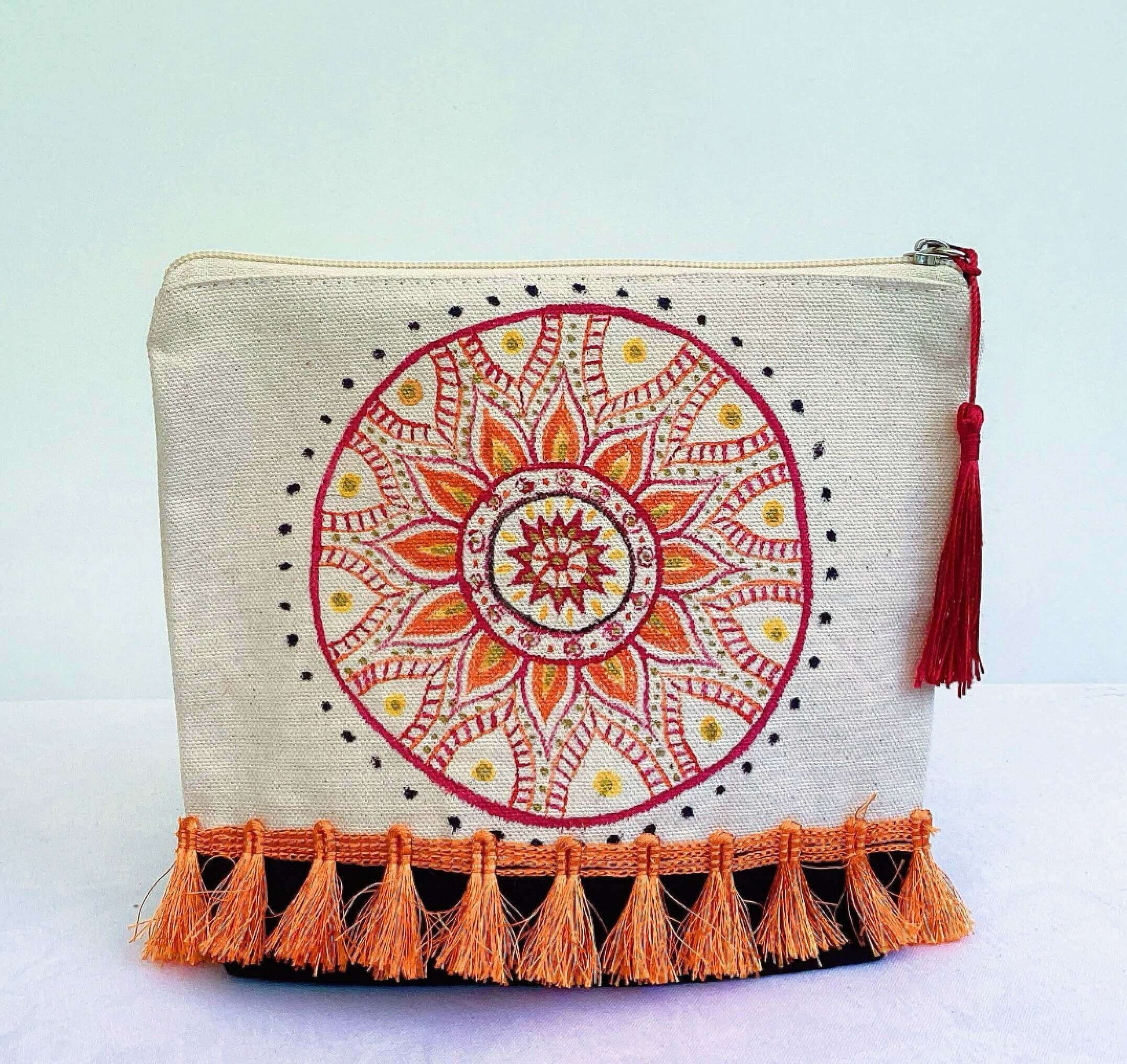 Front view of make-up pouch, black canvas base, with hand painted red and orange mandala, red tassel and zip, orange fringe.