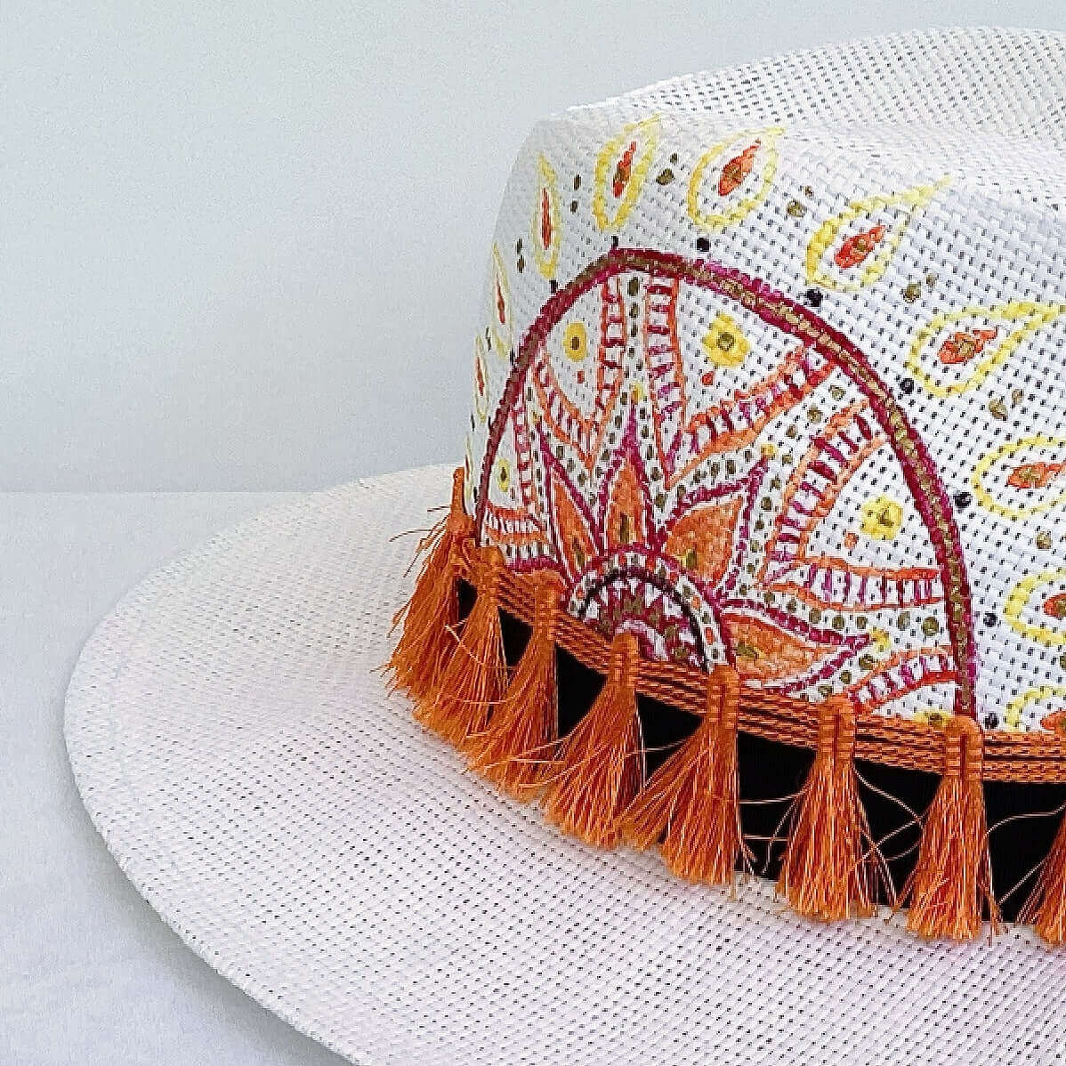 Close up view of panama hat, black band with orange fringe at base, and hand painted red, gold and orange mandala.