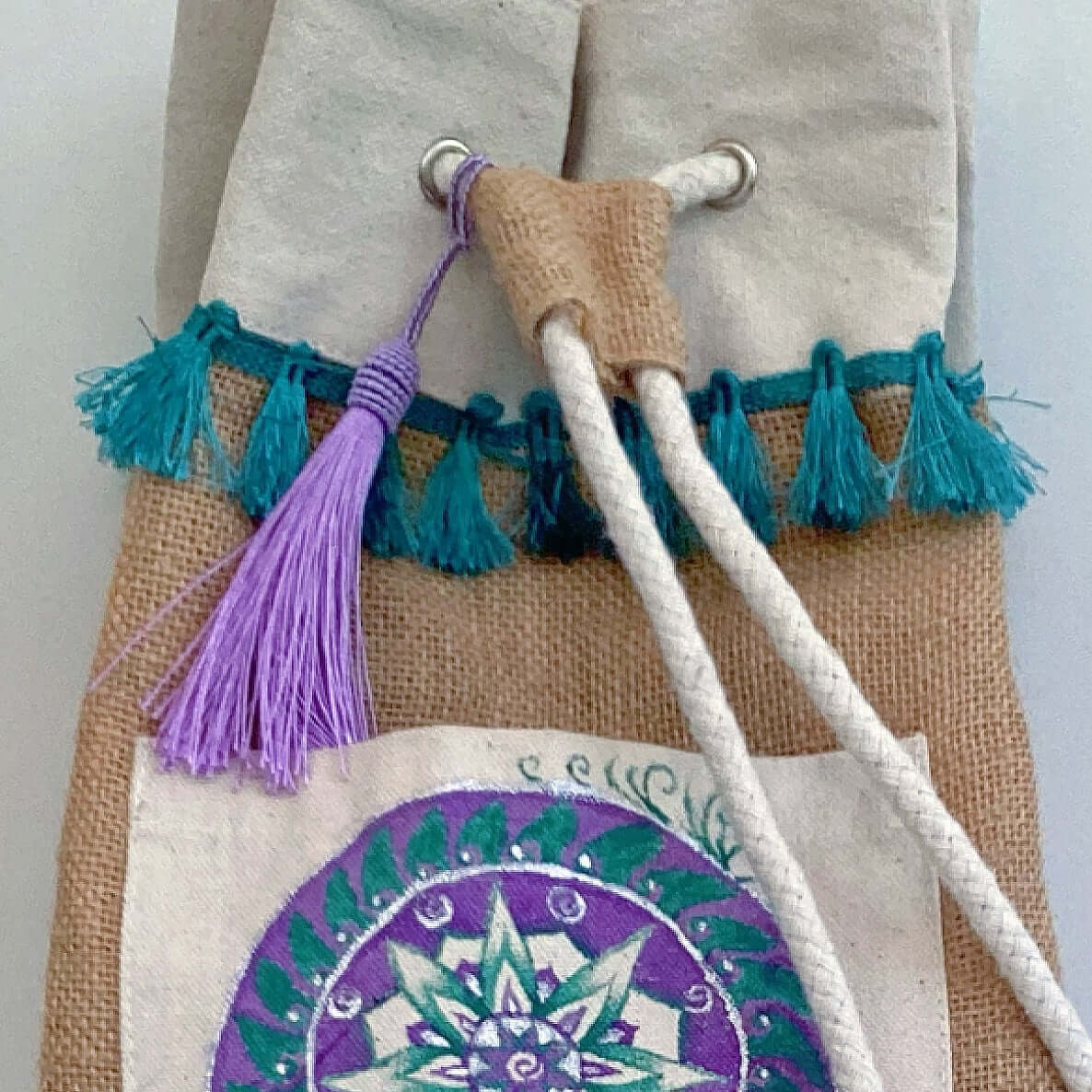 Detail view of hessian satchel with hand painted teal and purple mandala on canvas patch, with teal fringing at the top.