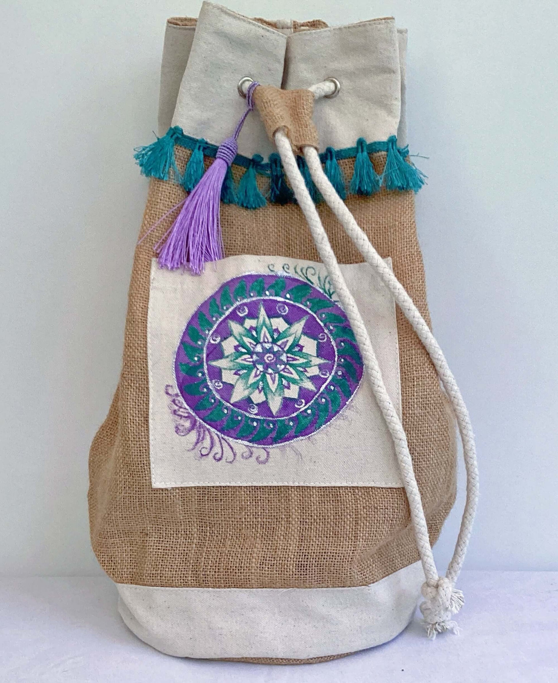 Front view of hessian satchel with hand painted teal and purple mandala on canvas patch, with teal fringing at the top.