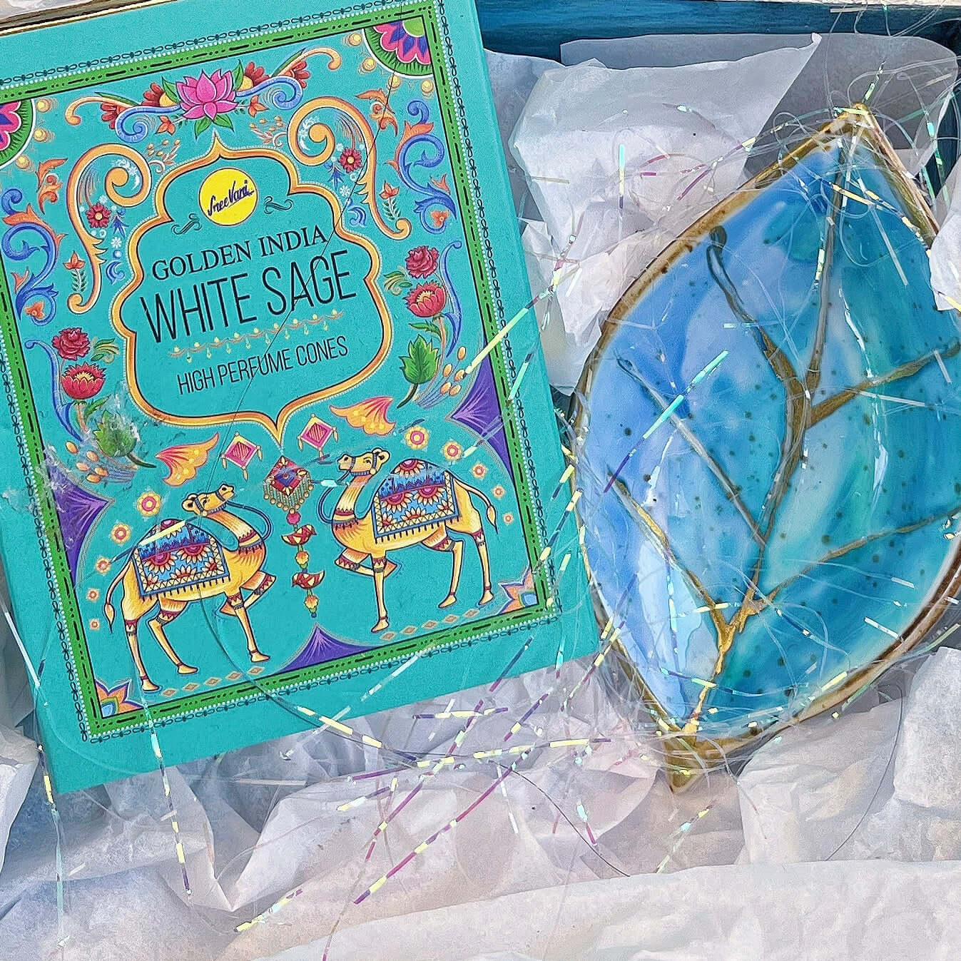 Contents detail view of incense and turquoise painted ceramic leaf, nestled in white tissue paper and tinsel.
