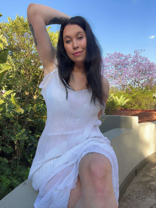 Front view of model sitting on a wall wearing the white chiffon two piece, trees and sky in the background.