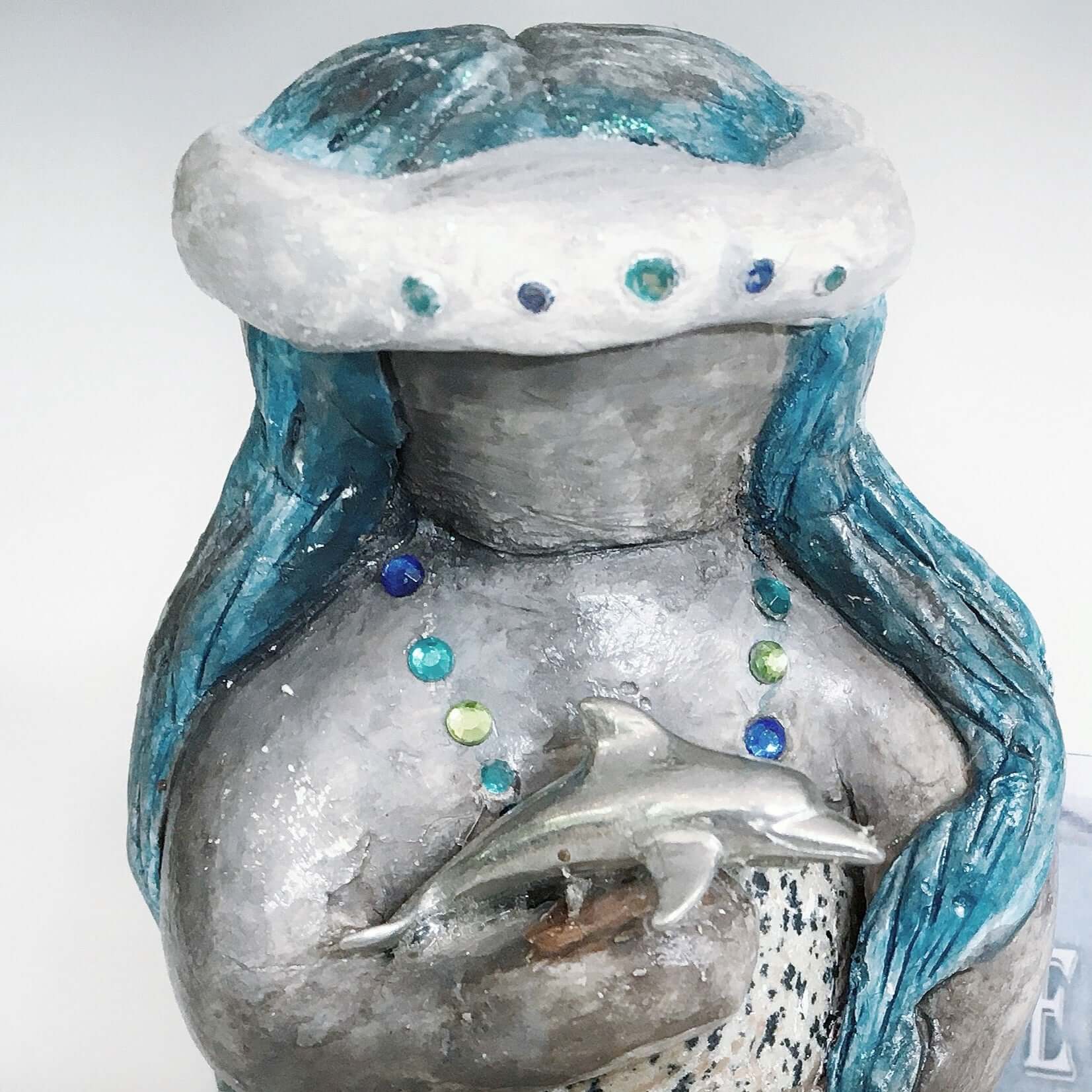 Close up of elemental, brown wash painted figure, jewel band on head, bluish long hair, holding a stone and dolphin.
