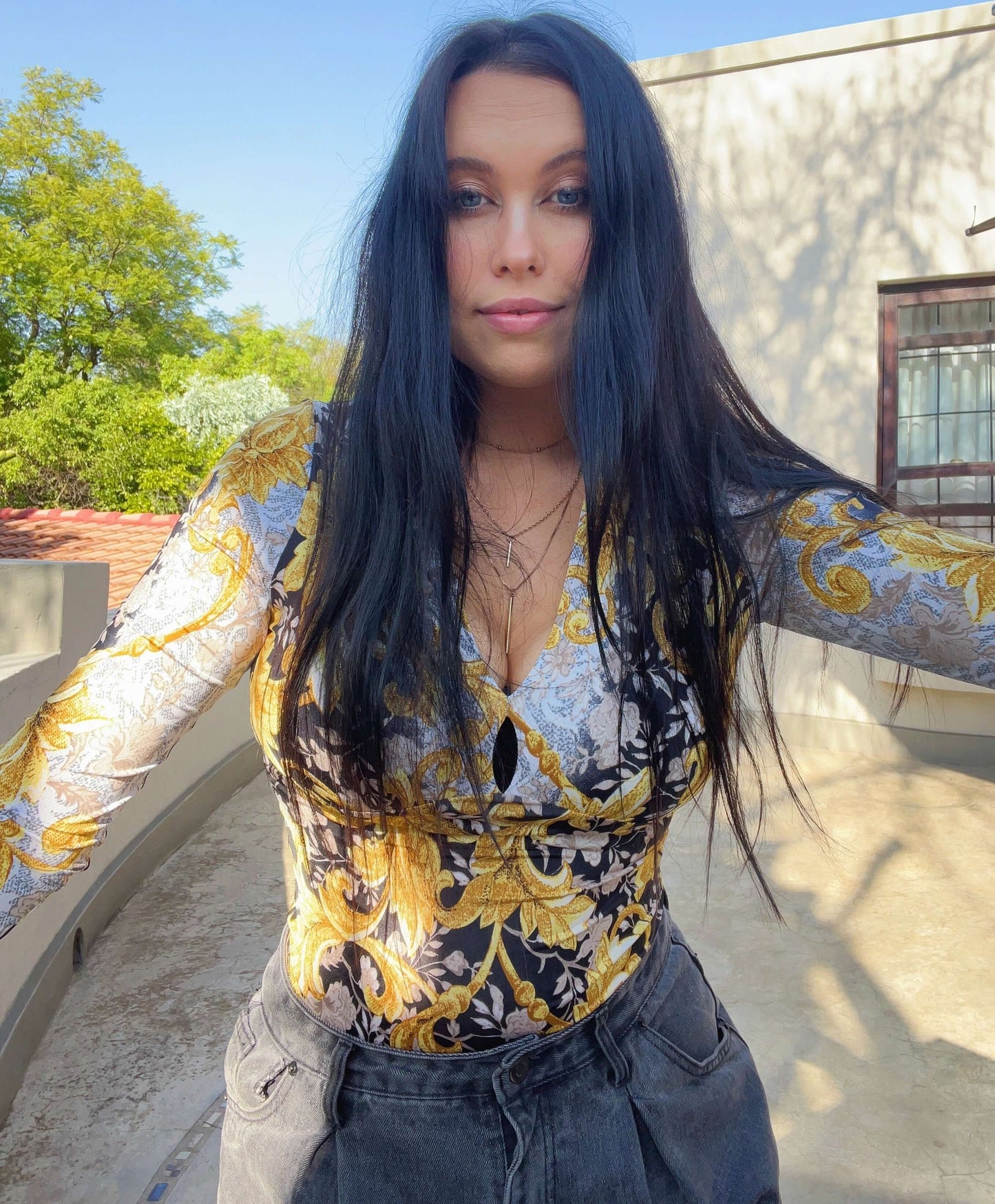 Front view of top, model is wearing black jeans, her hands outstretched, palm trees in the background.