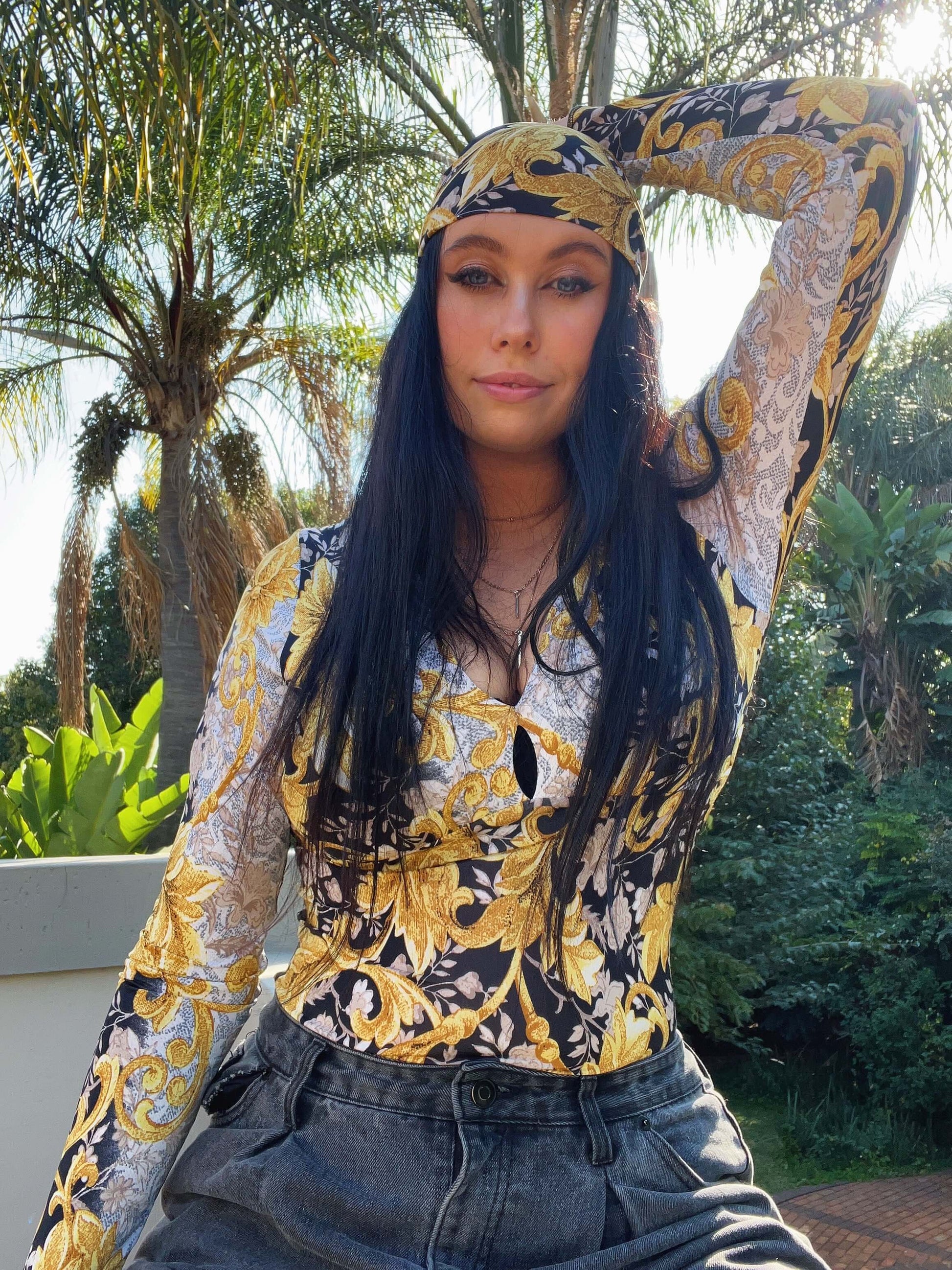 Front view of top, model is sitting on a low wall, is wearing a matching fabric bandana, palm trees in the background.