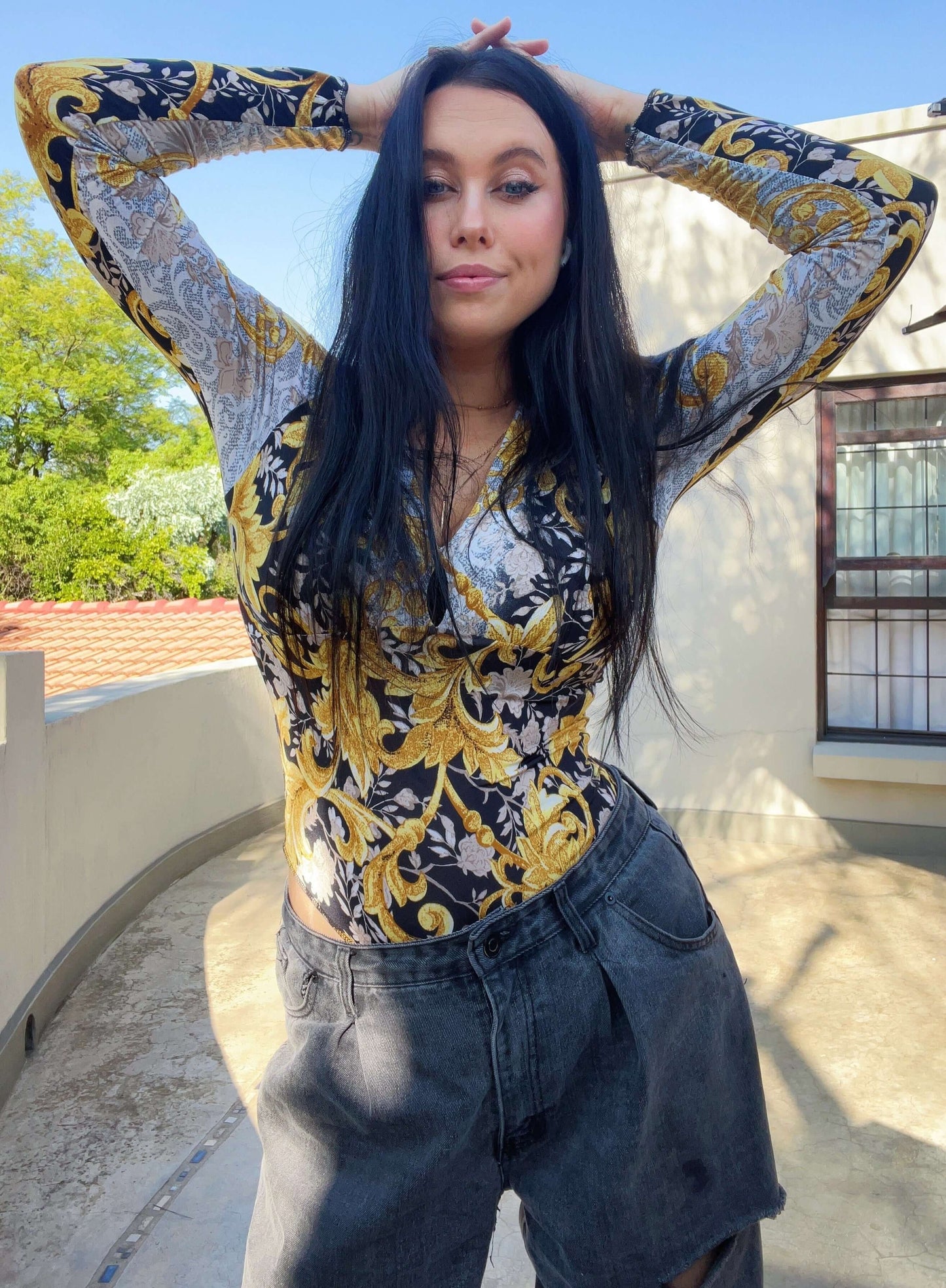 Front view of top, model is wearing black jeans, her hands on her head, palm trees in the background.