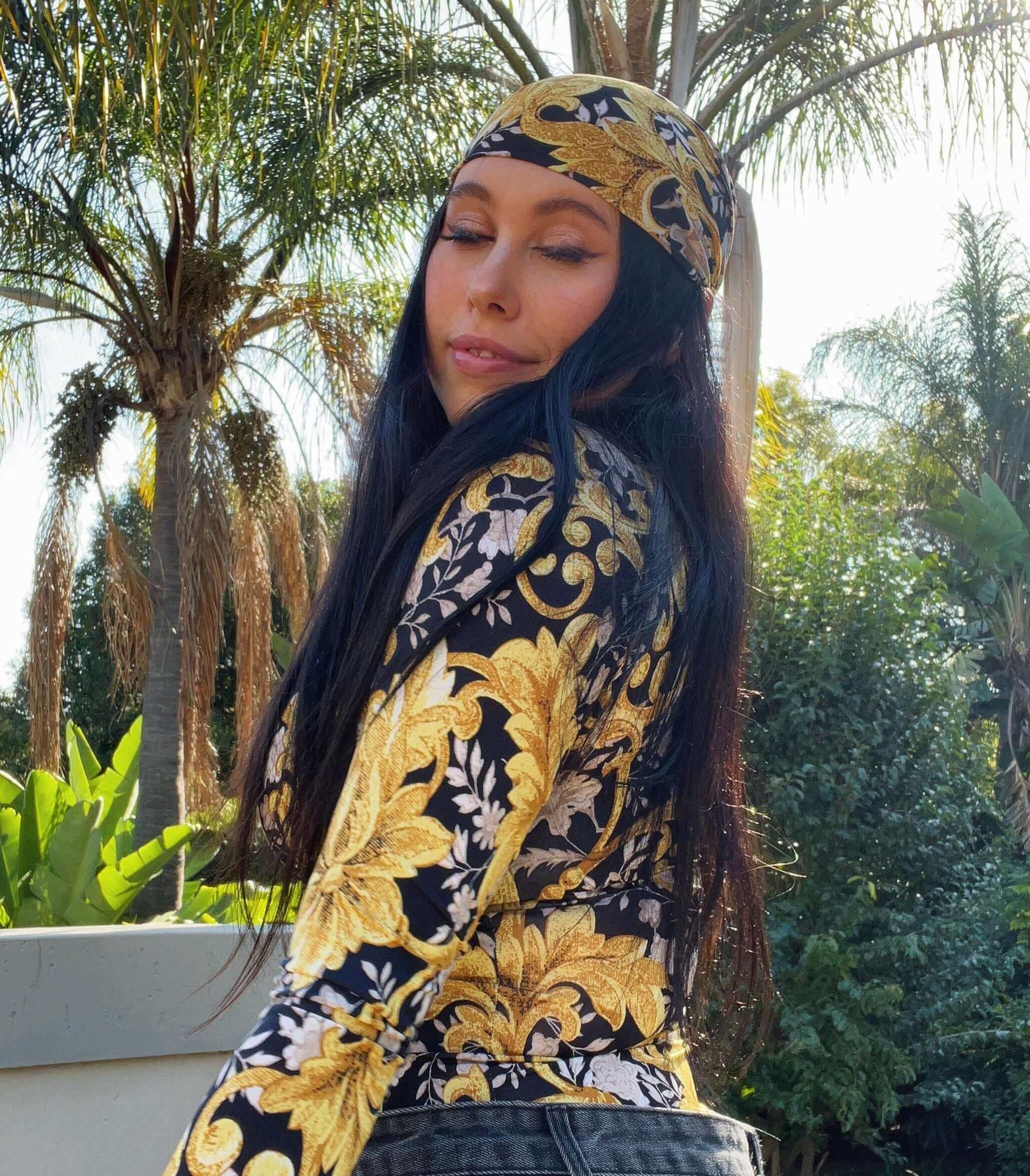 Side view of top, model is leaning into a low wall, is wearing a matching fabric bandana, palm trees in the background.