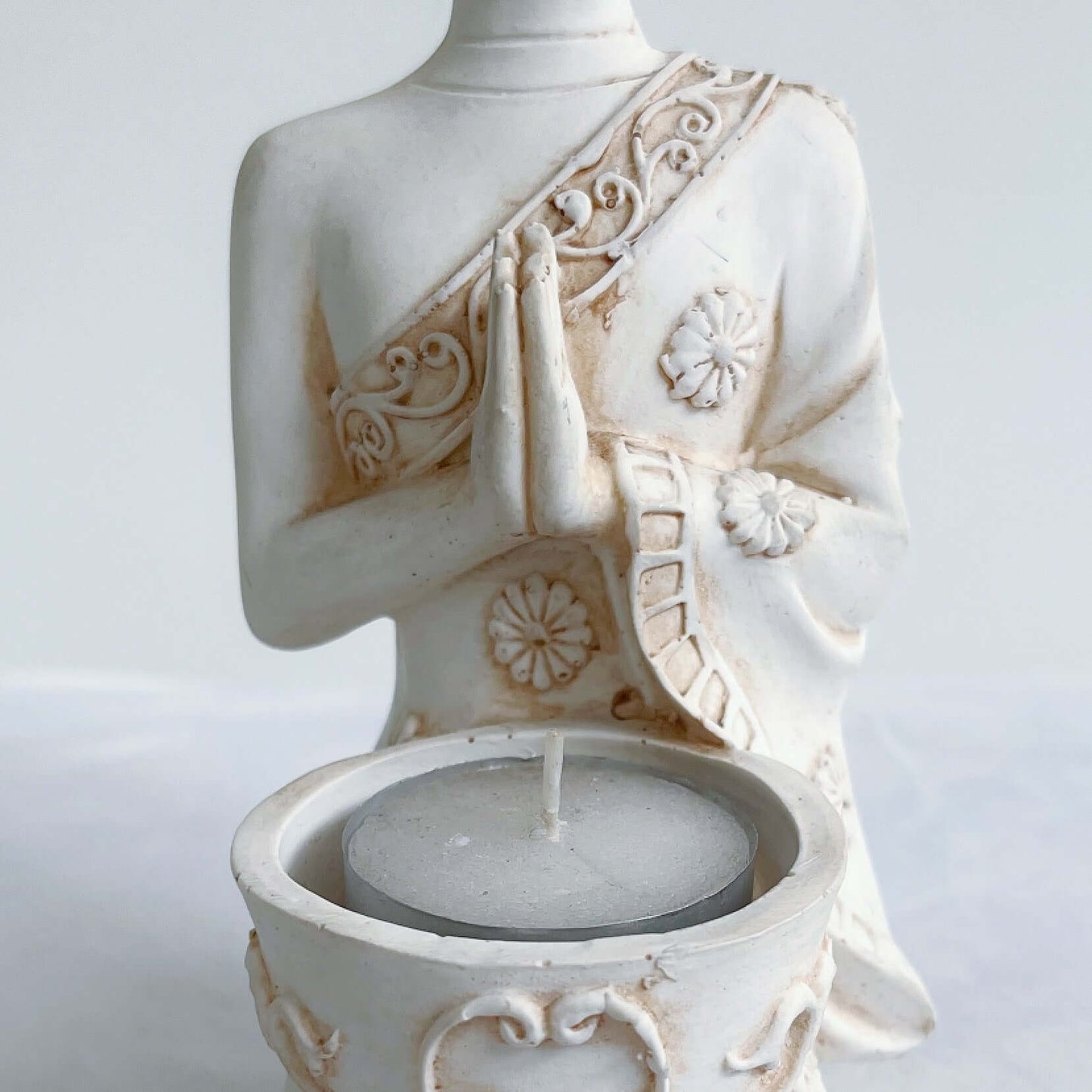 Detail view of buddha kneeling, candle holder on lap, with ornate robes and detailing, natural off white colors.