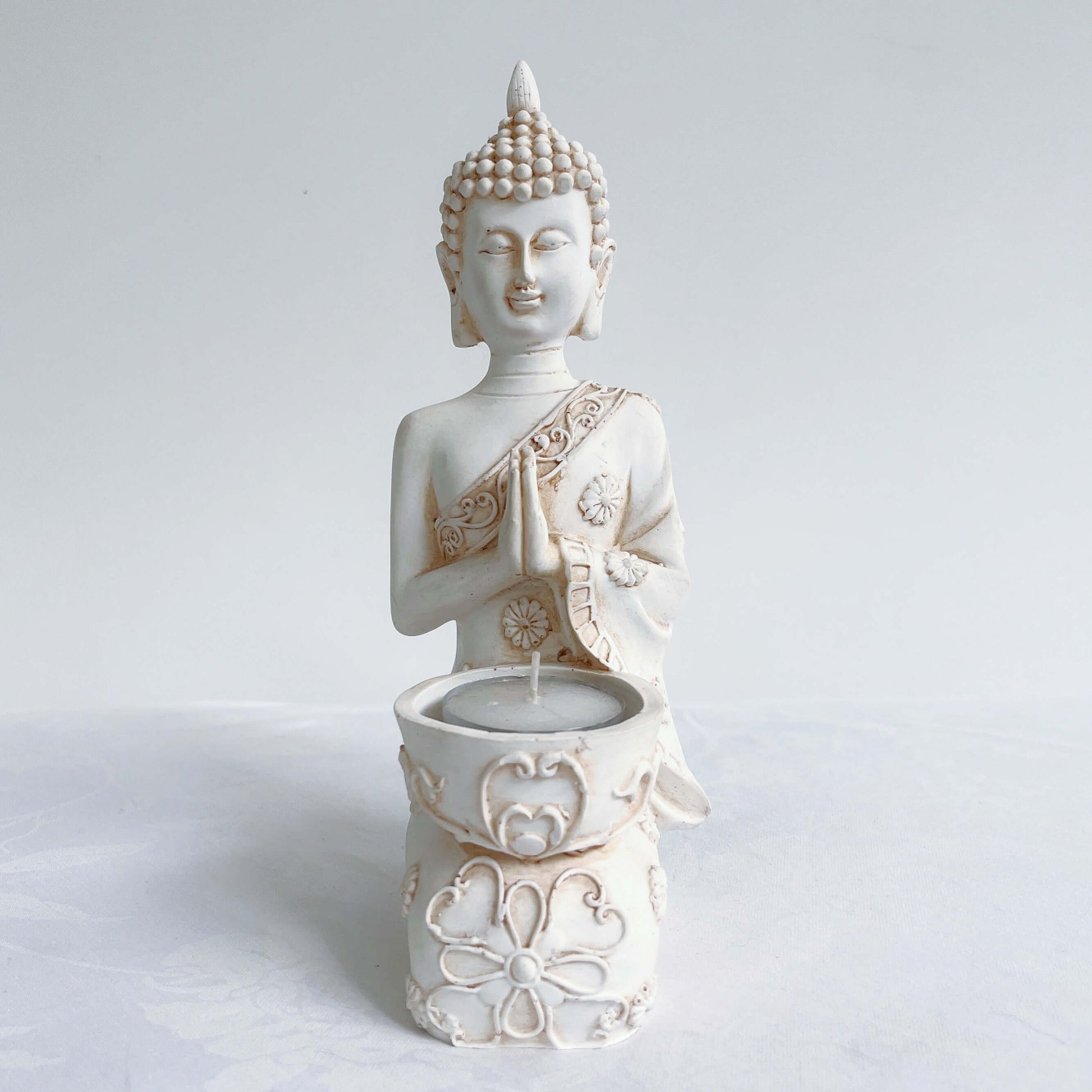 Front view of buddha kneeling, candle holder on lap, with ornate robes and detailing, natural off white colors.