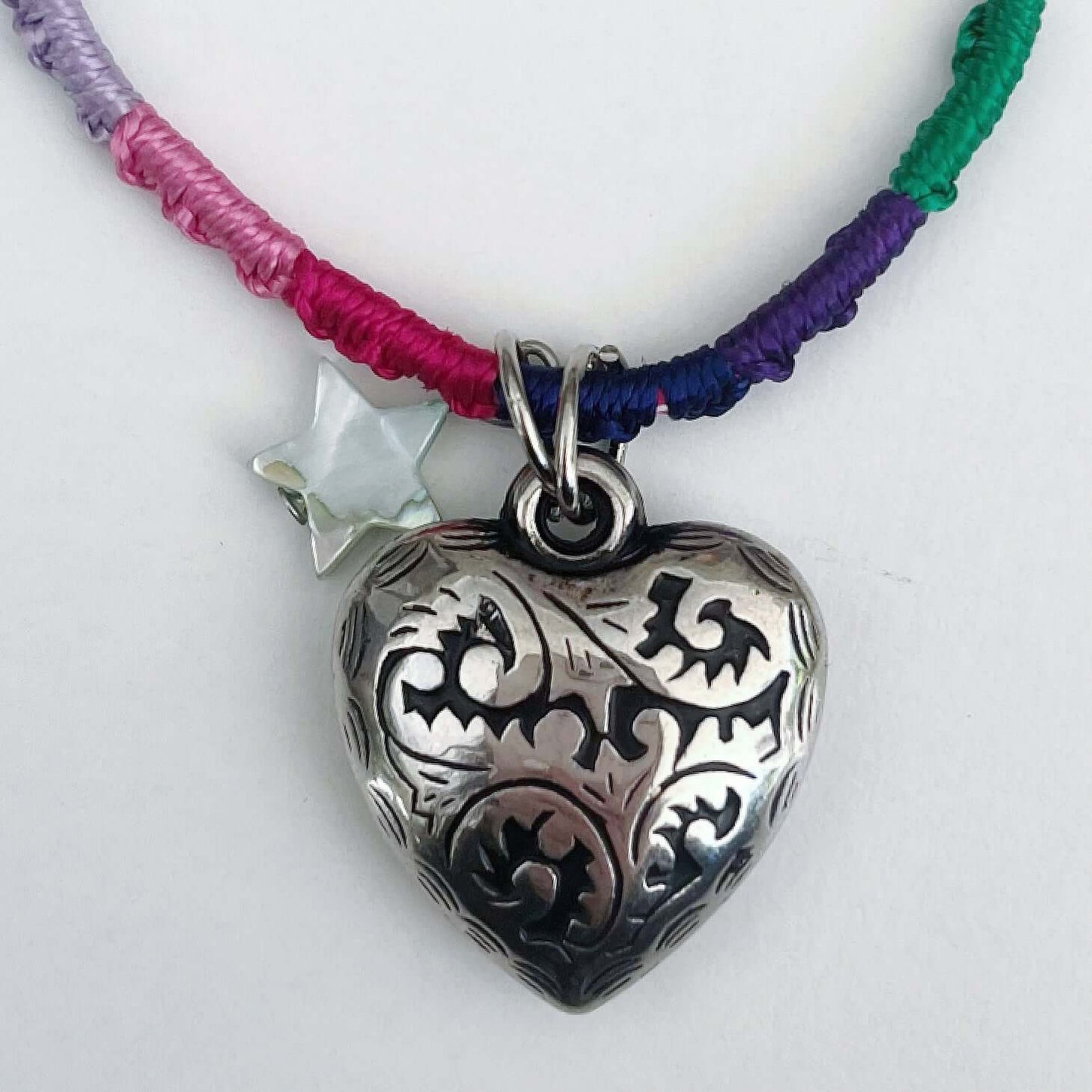 Detail of silver metal filigree heart 25mm, little mother of pearl star, on silky multi colored, wound and knotted thread.