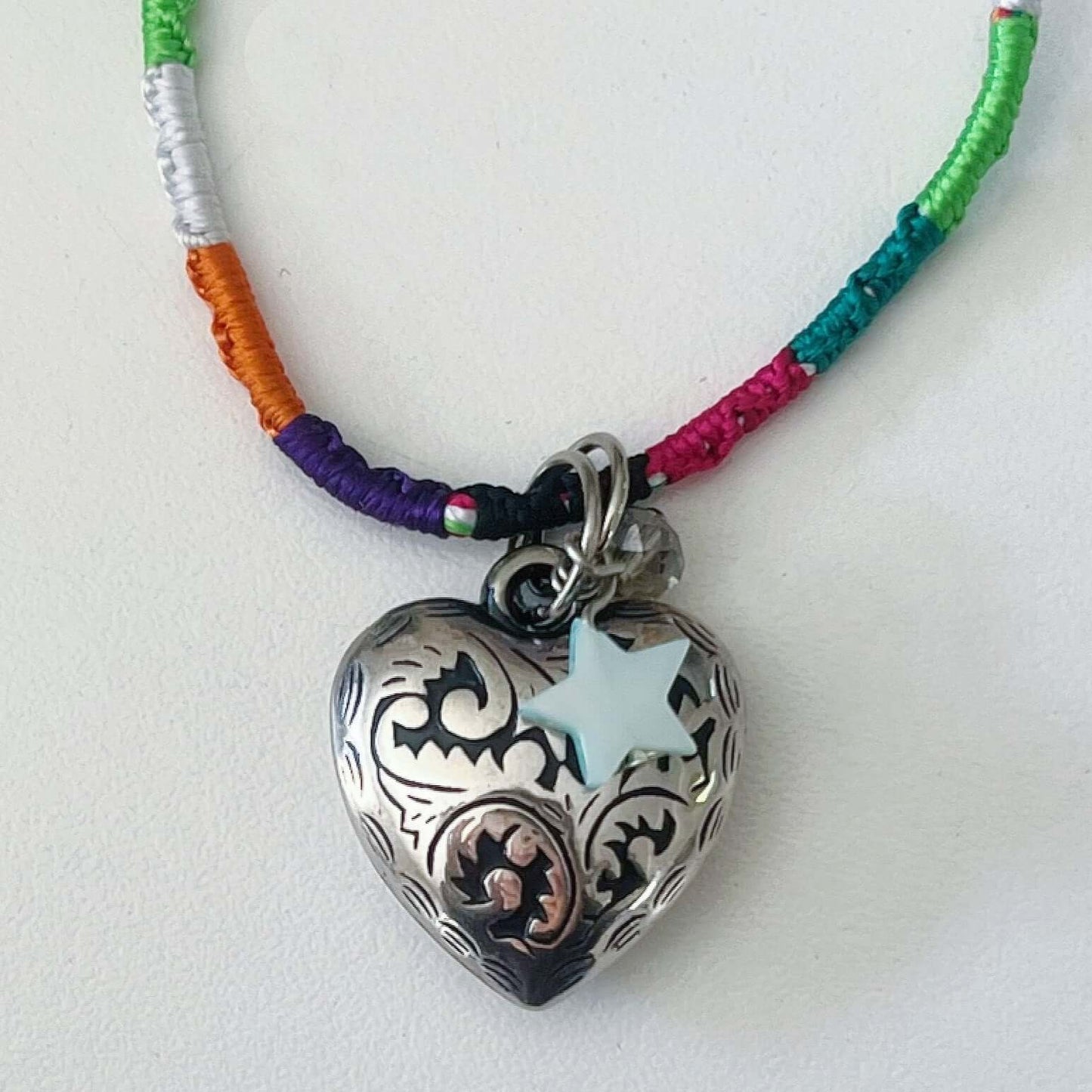 Detail of silver metal filigree heart 25mm, little mother of pearl star, on silky multi colored, wound and knotted thread.