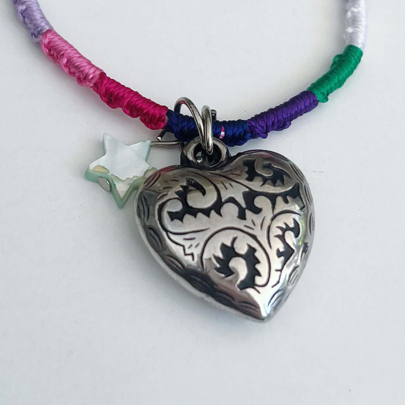 Detail of silver metal filigree heart 25mm, little mother of pearl star, on silky multi colored, wound and knotted thread.