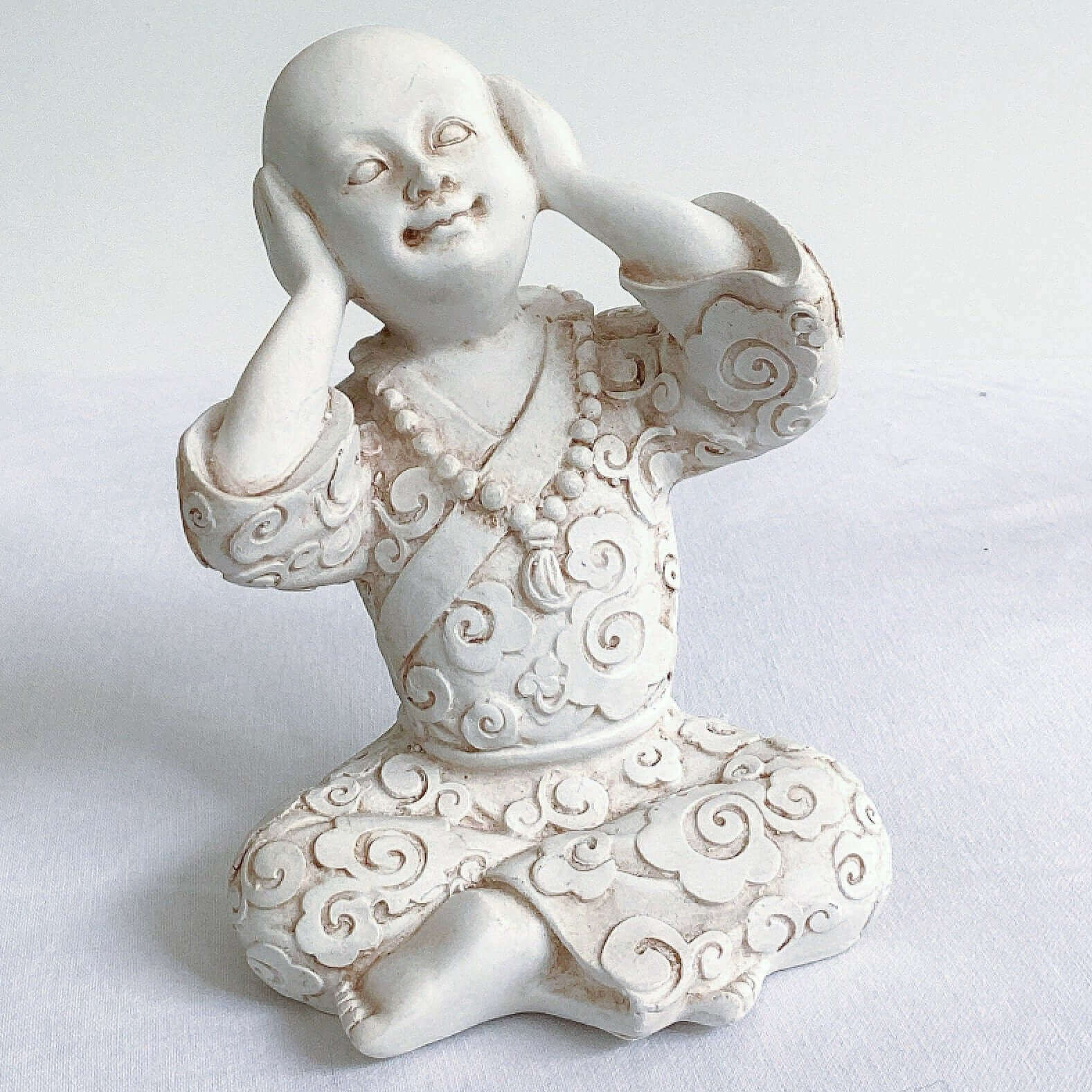 View of single monk, hear no evil, natural off white colors, with ornate robes, and hands covering the ears.