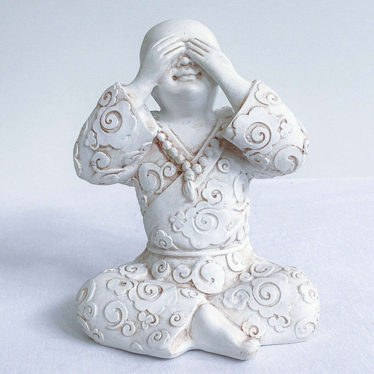 View of single monk, see no evil, natural off white colors, with ornate robes, and hands covering the eyes.