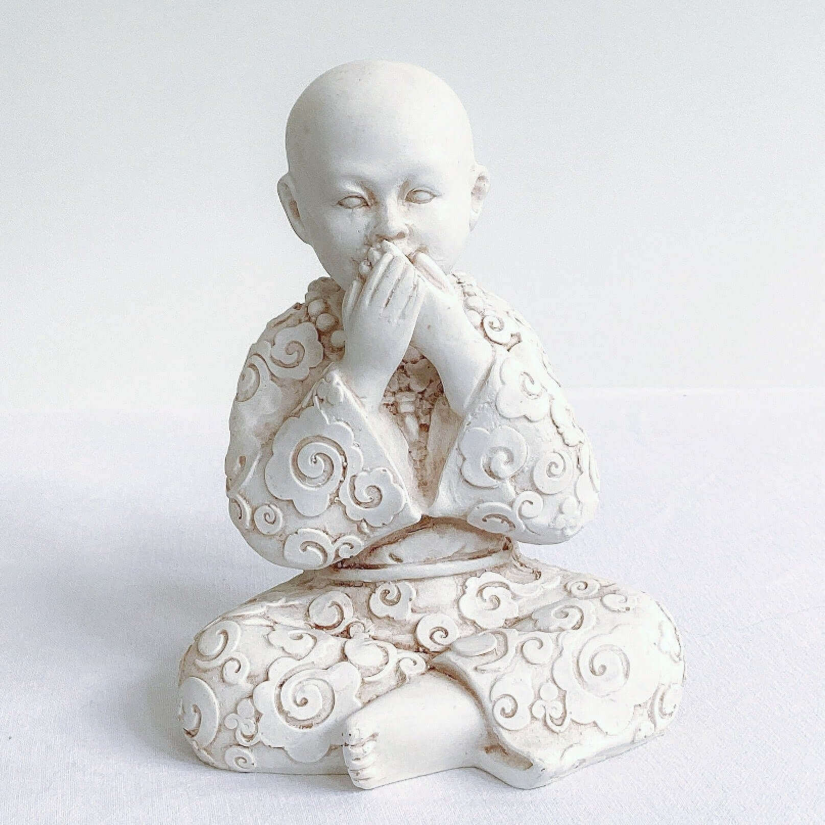 View of single monk, speak no evil, natural off white colors, with ornate robes, and hands covering the mouth.