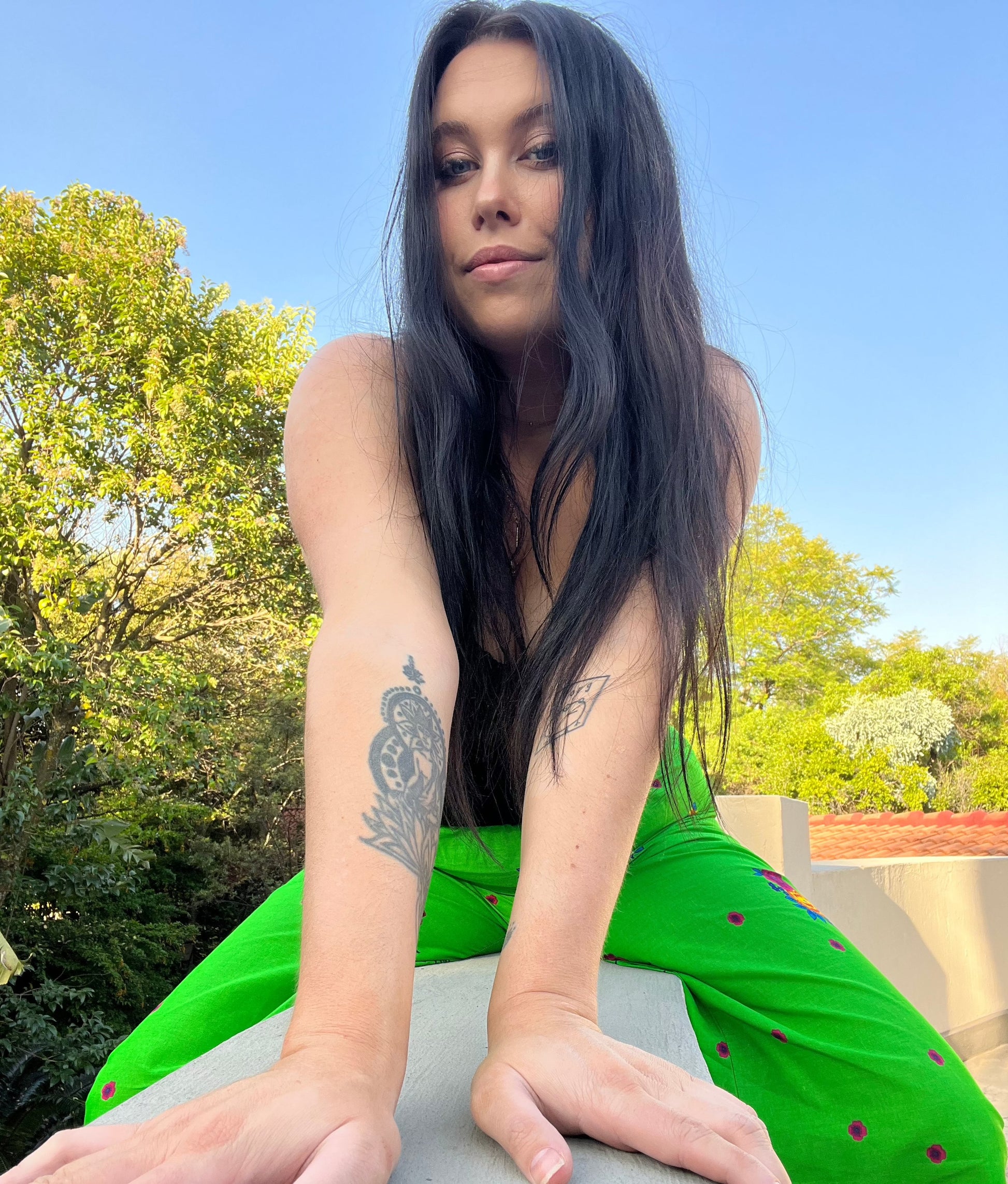 View of model in the pants sitting on a wall with her legs dangling down on either side. Trees and sky in the background.