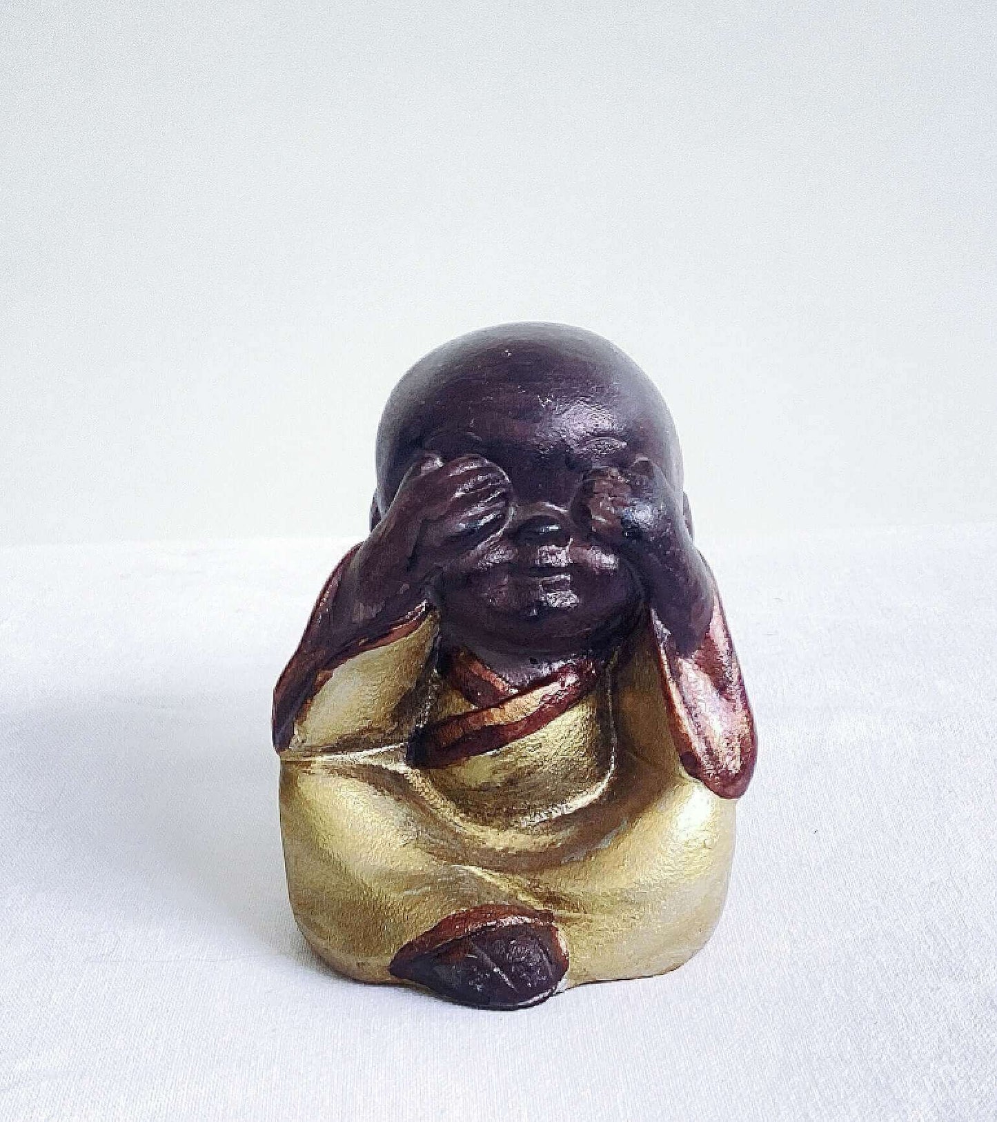 View of single monk, speak no evil, painted brown figure, with golden robes, and hands covering the mouth.