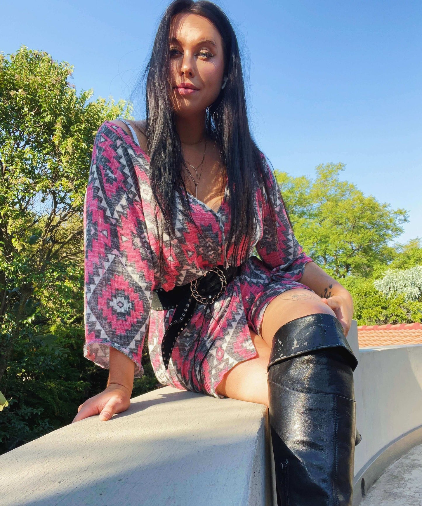 Model sitting on a wall wearing the dress and thigh-high boots, trees and sky in the background.