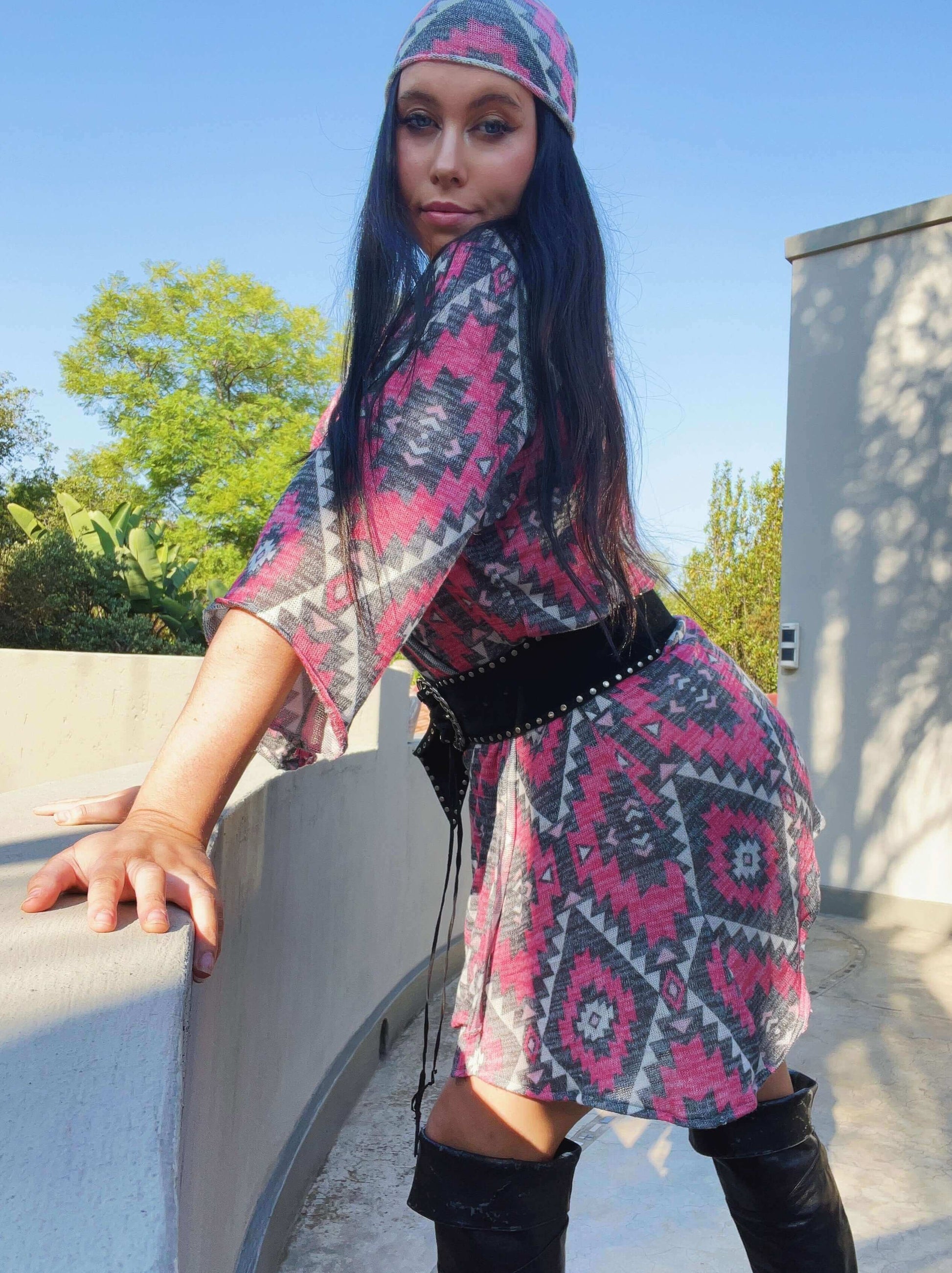 Model leaning her hands on a low wall, dress is with black belt, and thigh-high black boots, trees and sky in background.