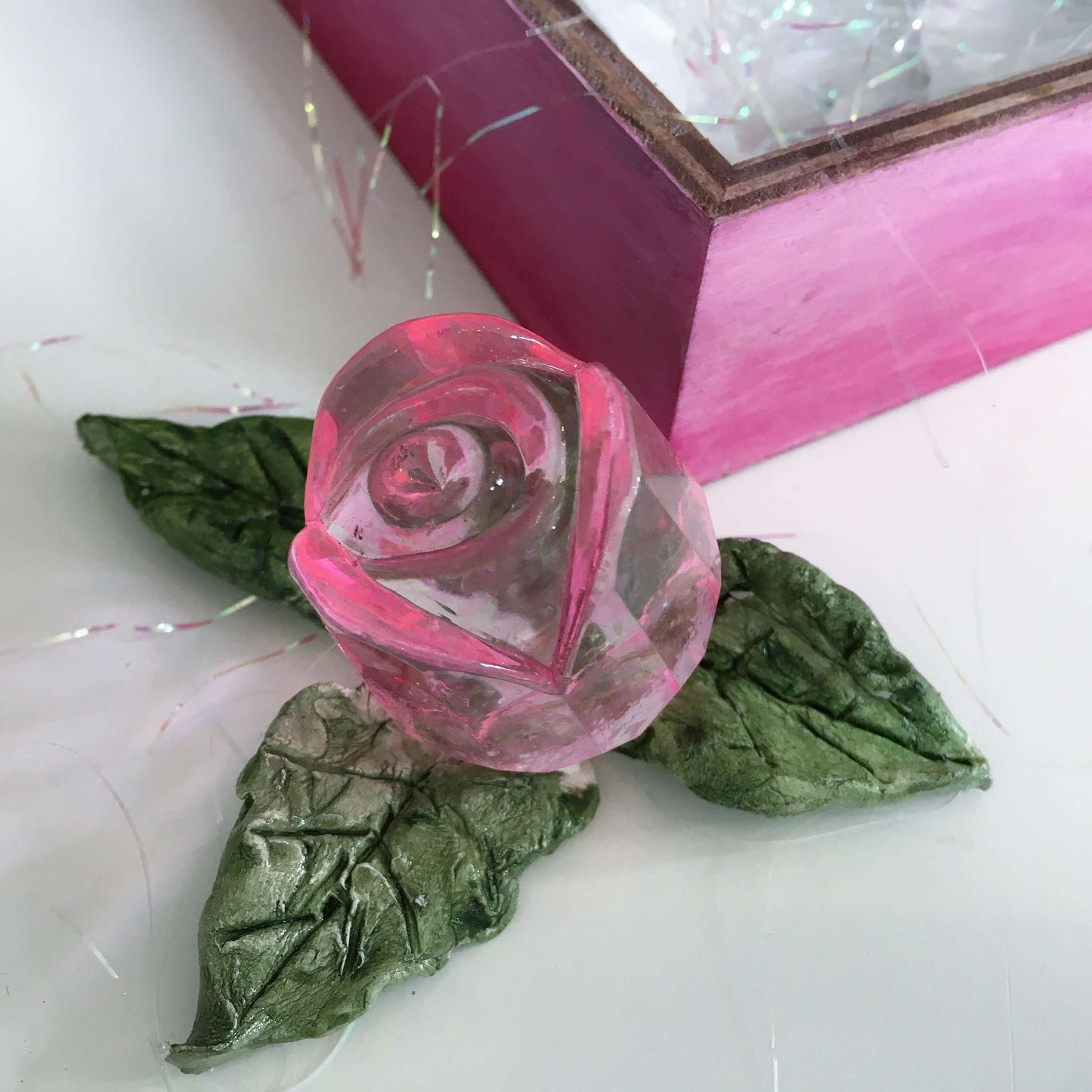 Detail view of pink glass rosebud and handmade ceramic leaves, next to its box.