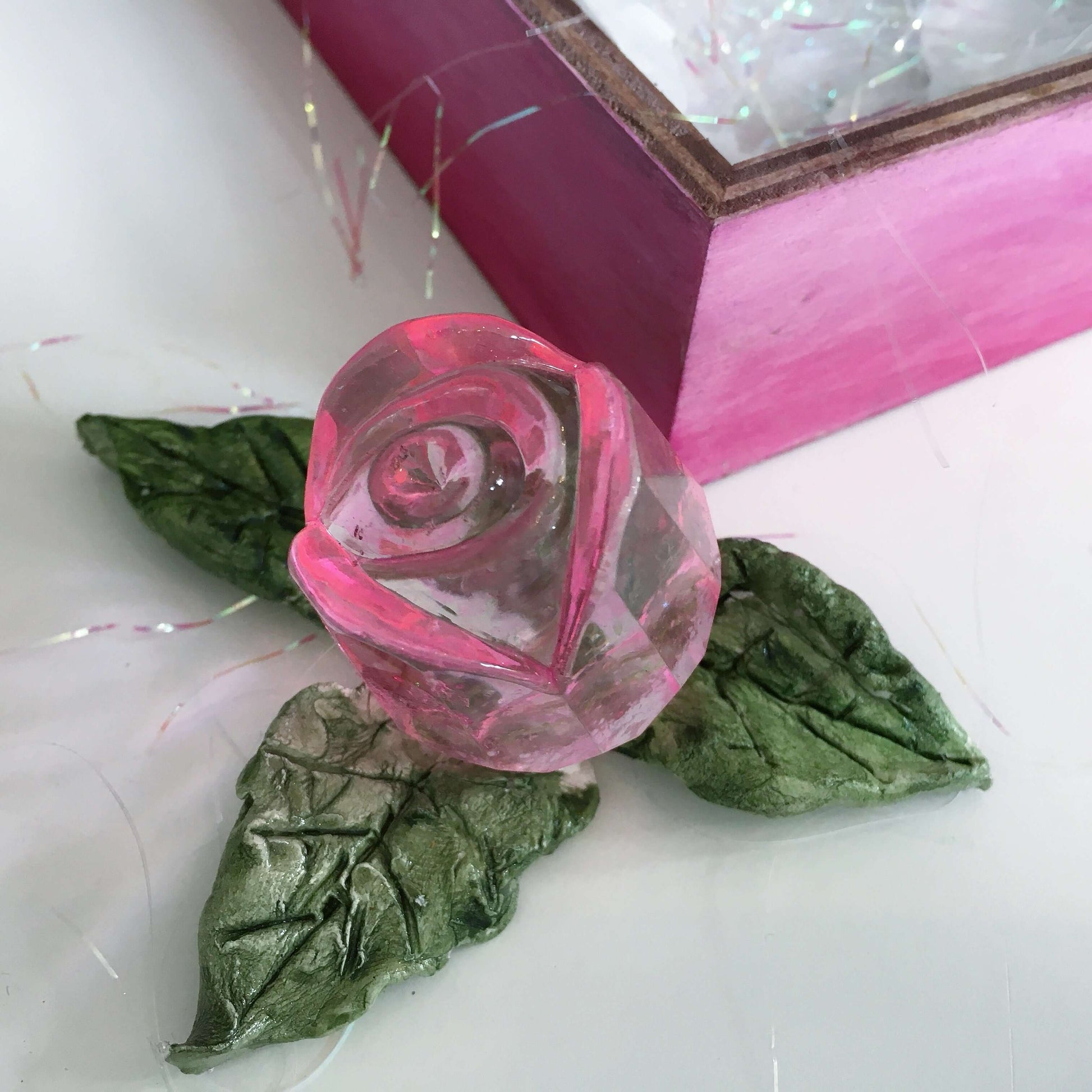 Detail view of pink glass rosebud and handmade ceramic leaves, next to its box.