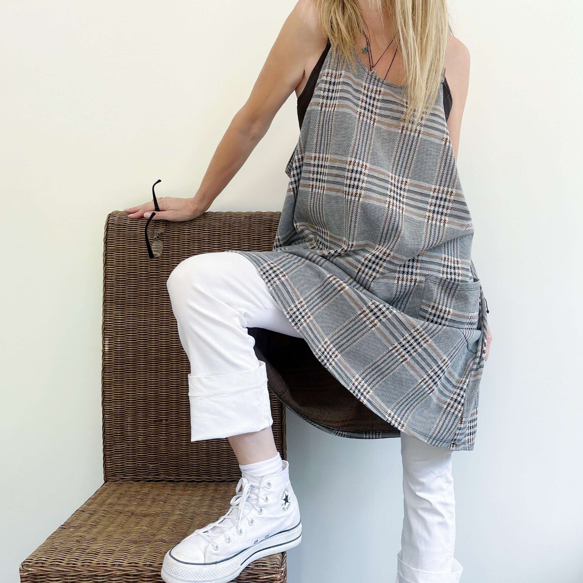 Front view of tartan dungaree dress, large pockets in front, model posing with a wicker chair.