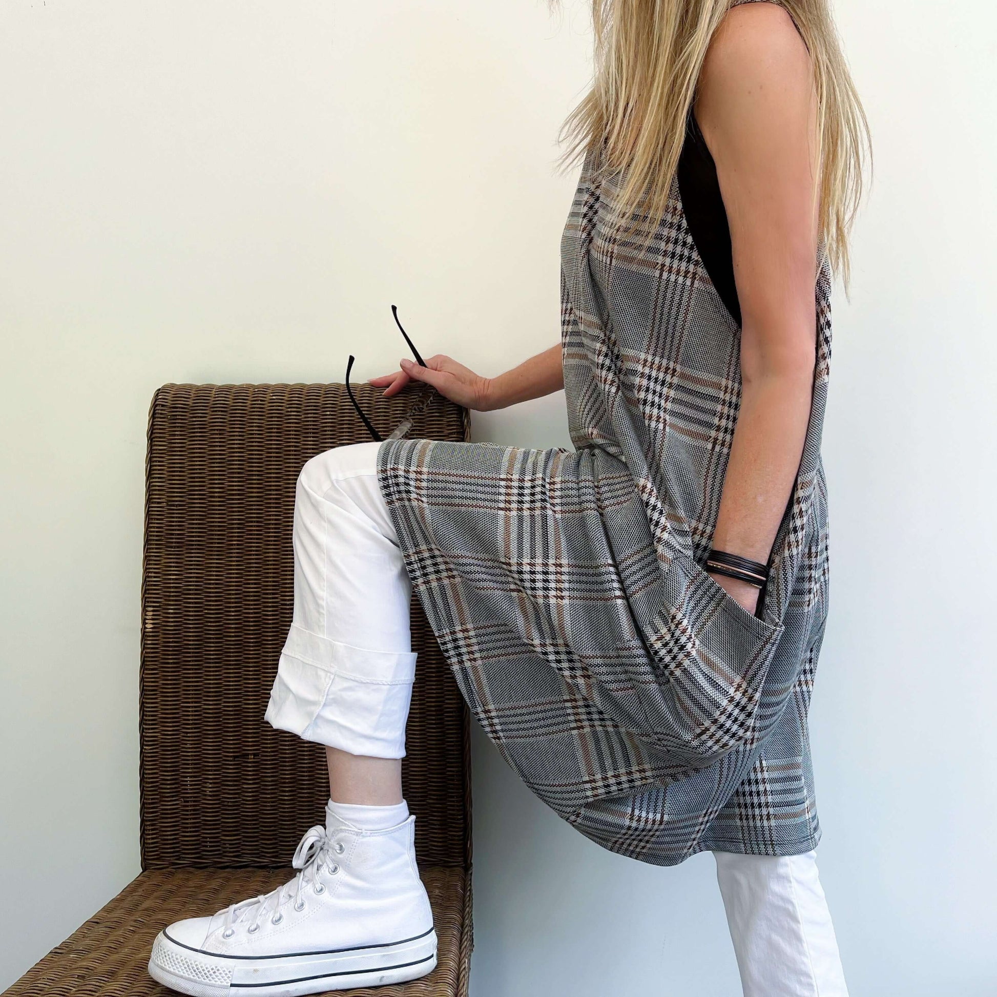 Side view of tartan dungaree dress, model sitting on a wicker chair, hands in pockets.