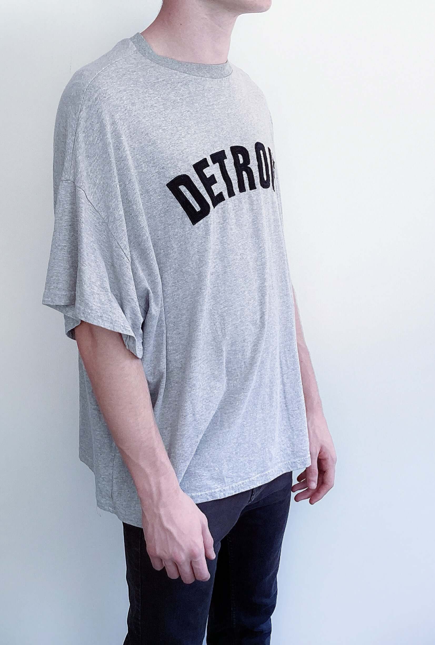 Side view of light grey, dropped shoulder tshirt, simple black bold printing, in an arc shape on the chest.