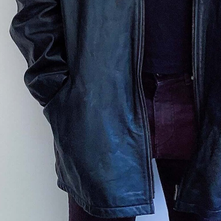 Front view of black leather jacket, mid thigh length, zip up, hands in pockets.