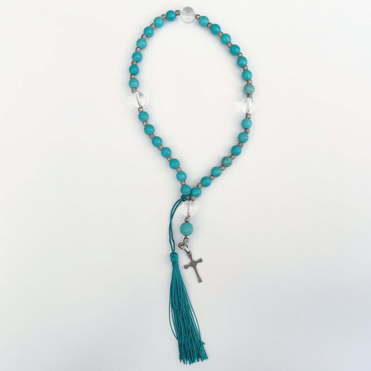 Top view of prayer beads, Blue Howlite sections, with one clear quartz here and there. Cross and tassel at the bottom.