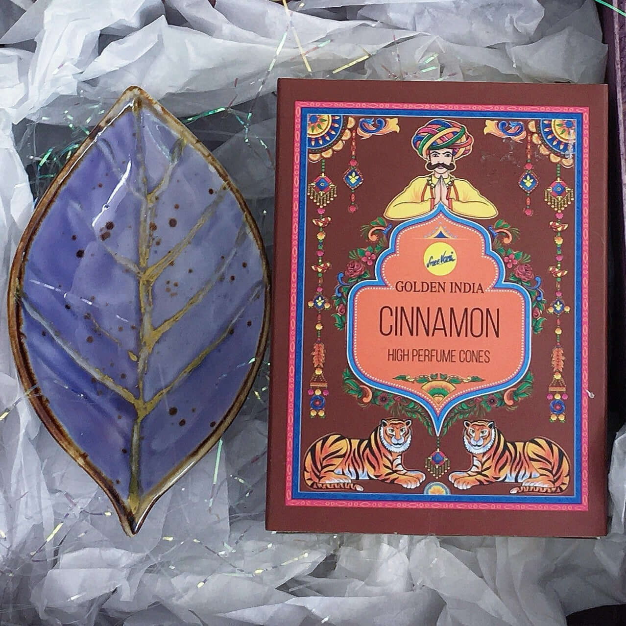 Contents detail view of incense and indigo painted ceramic leaf, nestled in white tissue paper and tinsel.