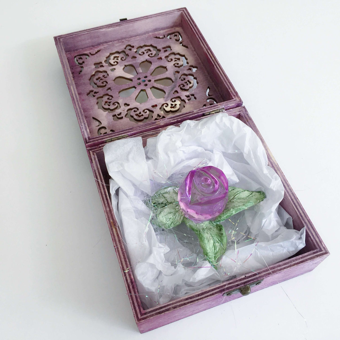 Contents of box, a purple glass rosebud with handmade ceramic leaves nestled in tissue paper and tinsel.